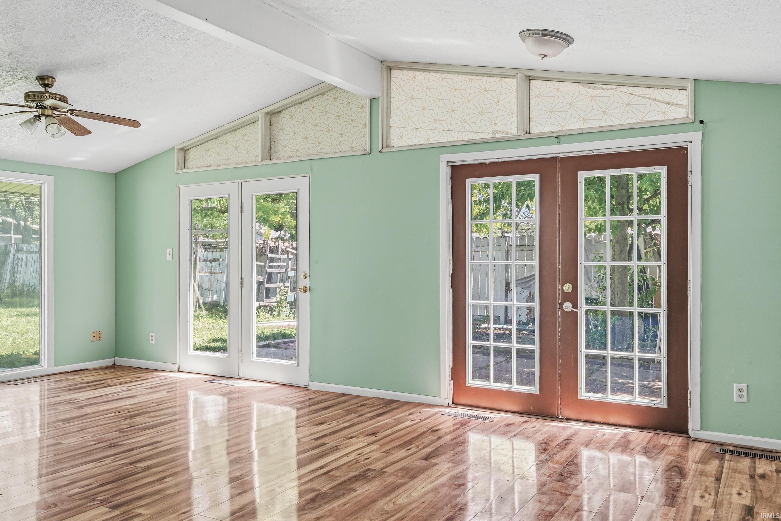 Vaulted ceiling with exposed beams, wood-finish flooring, two sets of French doors, and a ceiling fan with light fixture