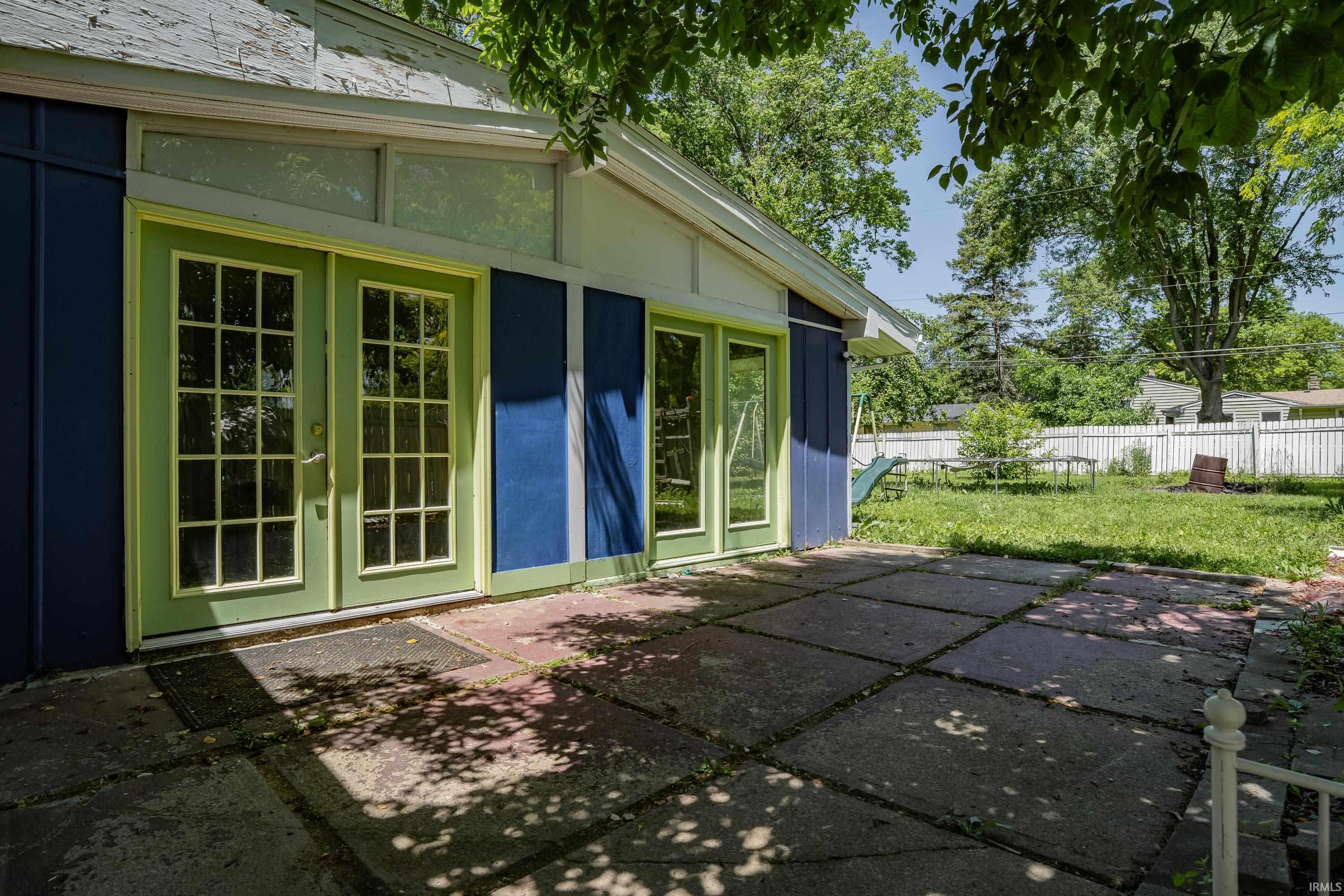 Rear exterior featuring a concrete block patio, light green French doors, blue siding, a white privacy fence, and mature trees