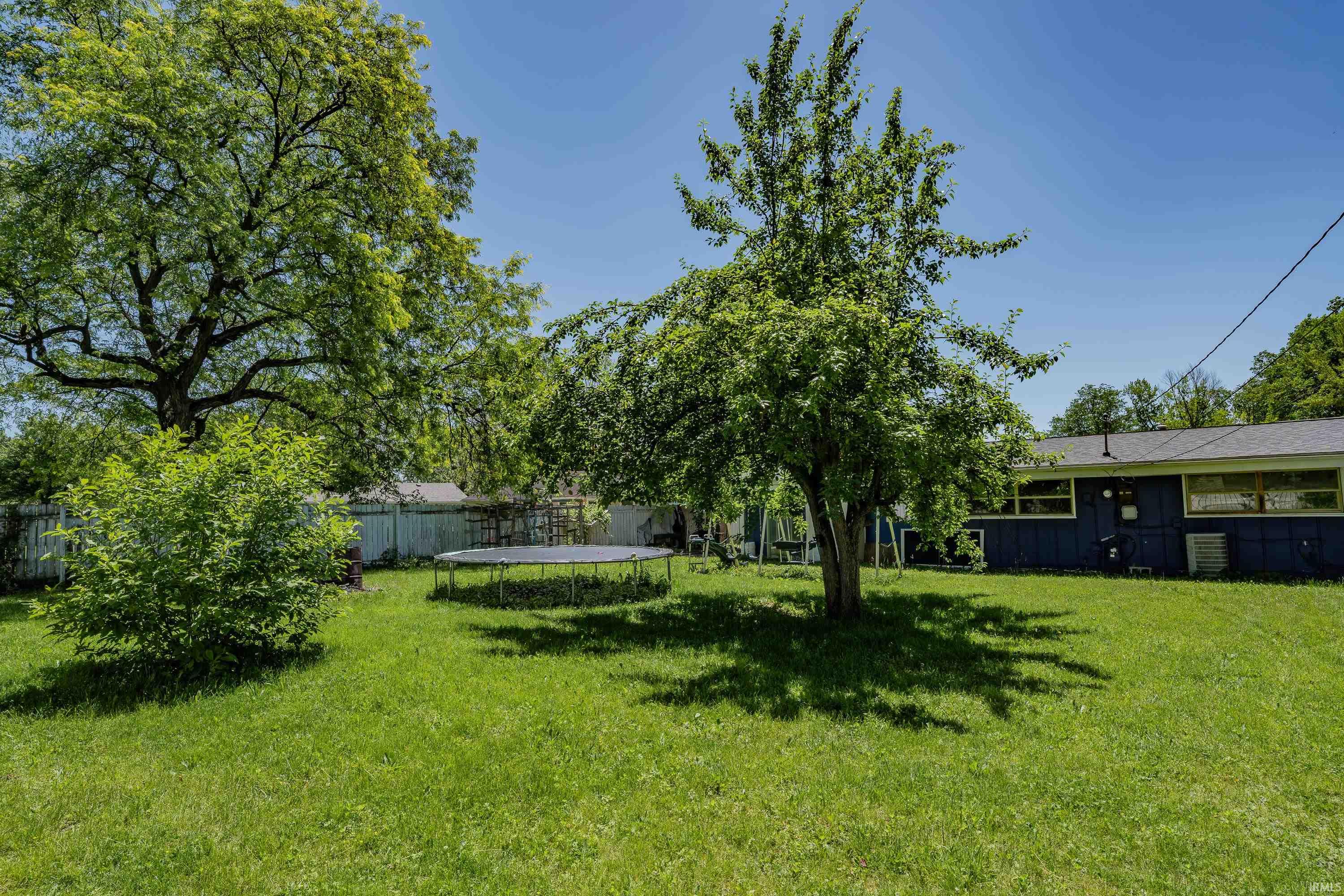Expansive grassy backyard featuring mature trees, a smaller ornamental tree, a trampoline, and a blue exterior home with multiple windows