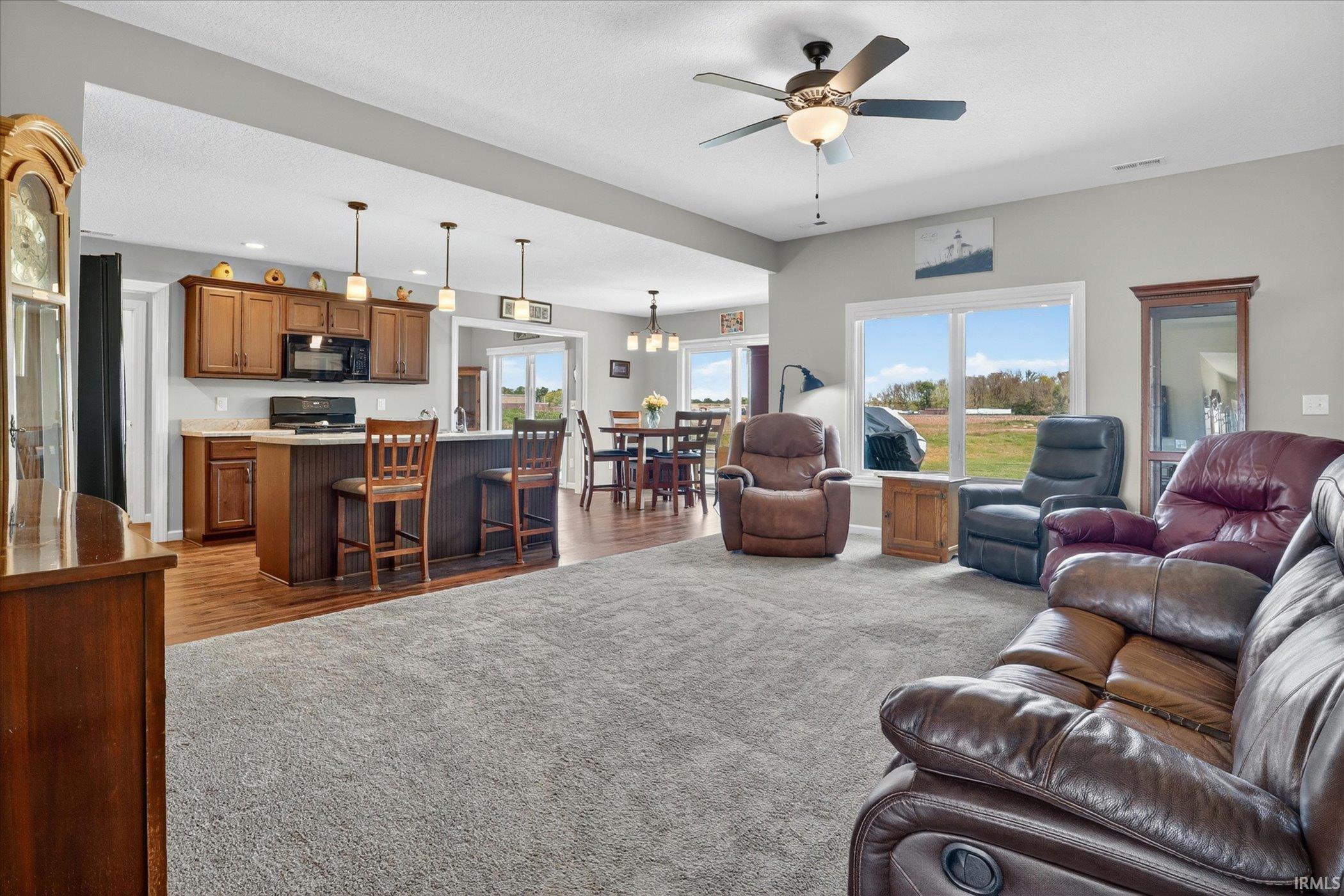 Open-concept living area featuring a spacious carpeted floor, an integrated kitchen with wood cabinetry and an island, a dining area with wood-finish flooring, and large windows with a rural landscape