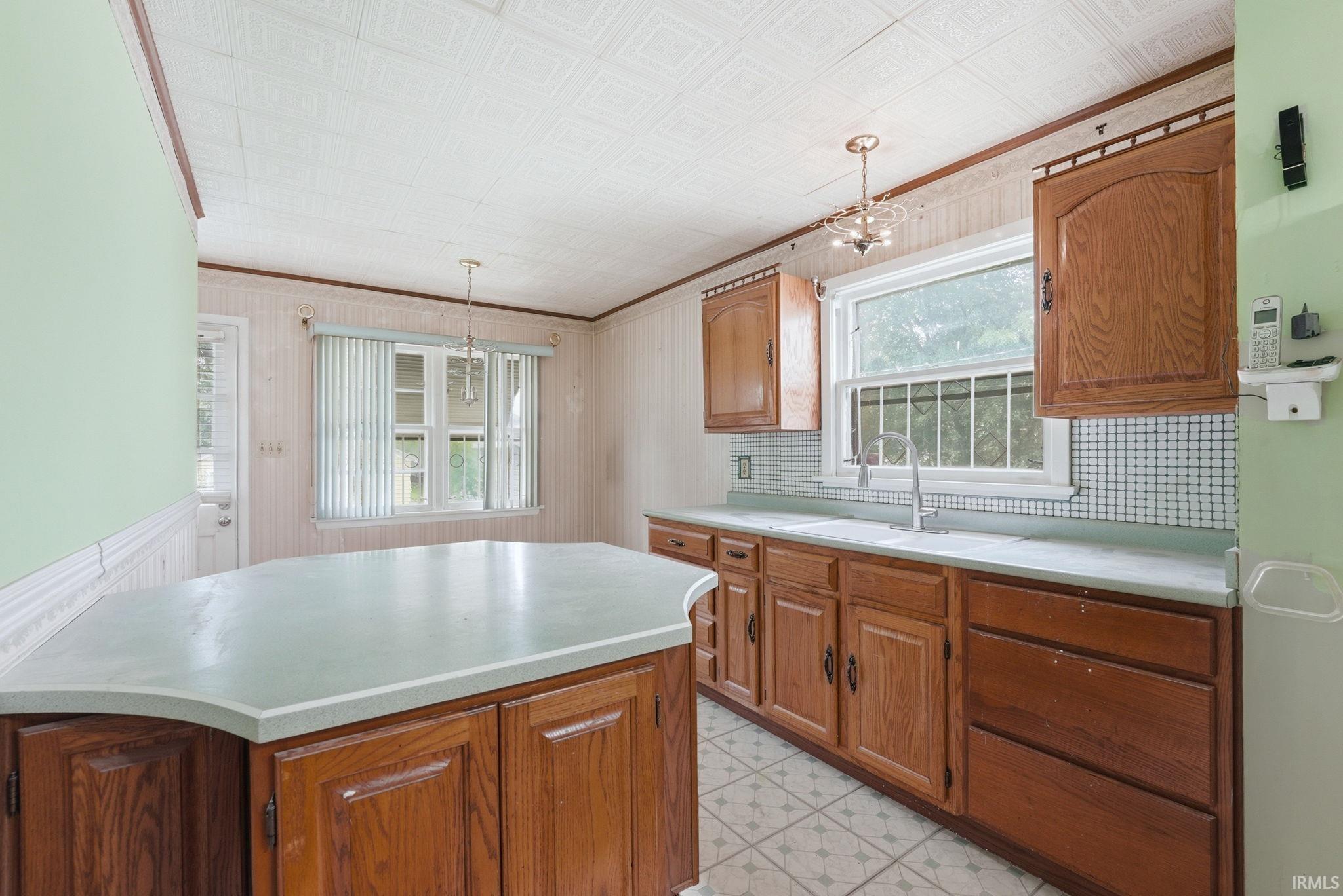 Kitchen featuring a central island with a light-toned countertop, wood-finish cabinetry, a large window over the sink, and patterned floor tiling