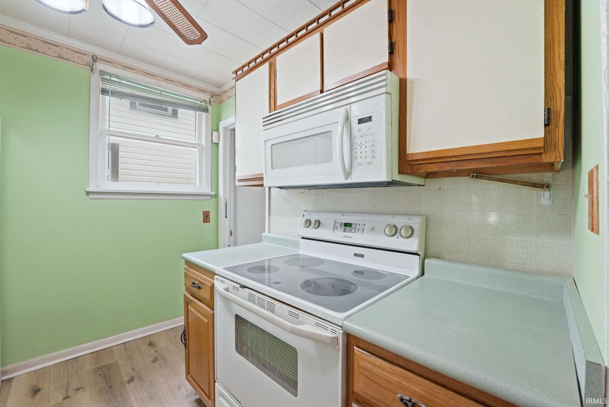 Kitchen featuring wood-finish flooring, light green walls, a window with blinds, wood cabinetry with white panels, and a white electric range with an overhead microwave