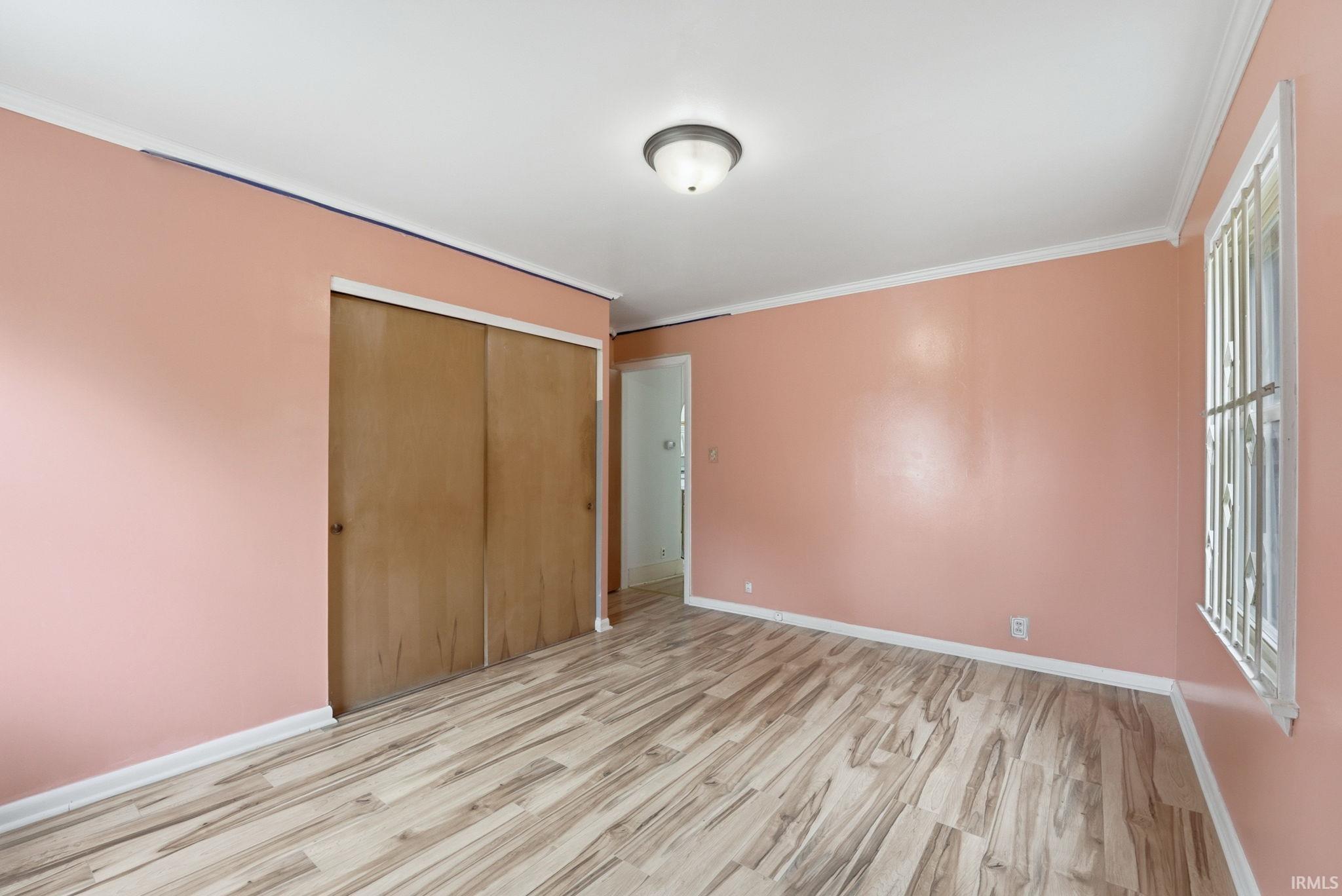 Spacious room featuring wood-finish flooring, sliding closet doors, a window with security bars, and a ceiling-mounted light fixture
