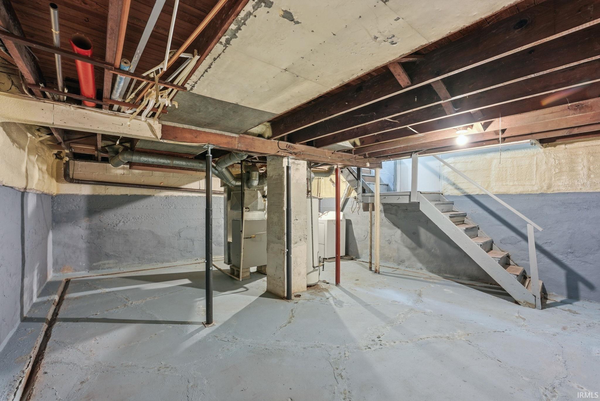 Unfinished basement with concrete flooring, exposed wood beam ceiling, painted concrete block walls, and a utility staircase