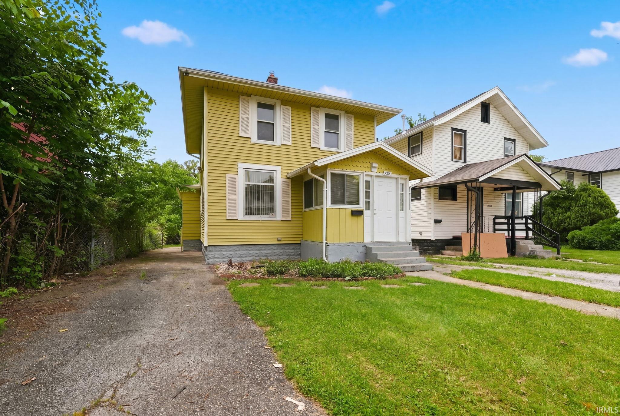 Two-story residence featuring light yellow siding with white window shutters, a covered porch entrance, and a paved driveway