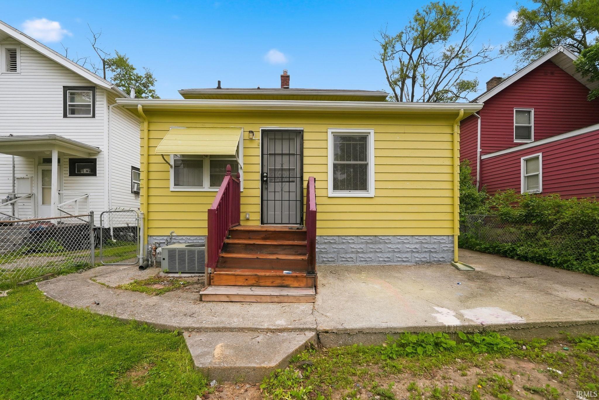 Rear exterior featuring yellow siding, a ground-level concrete patio, and wood stairs with a magenta railing