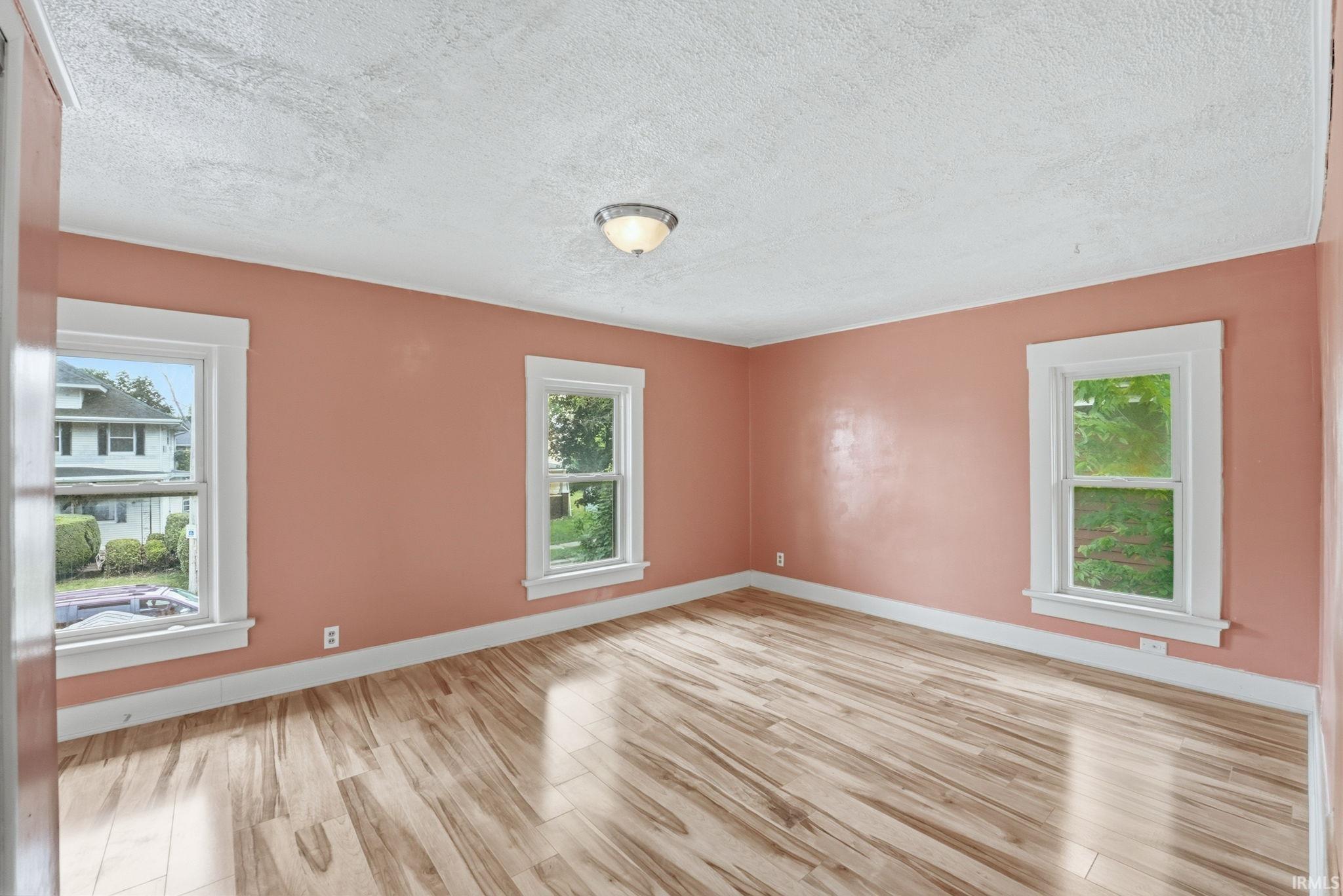 Open room featuring light wood-finish flooring, three windows with white trim, a textured ceiling, and a flush mount light fixture