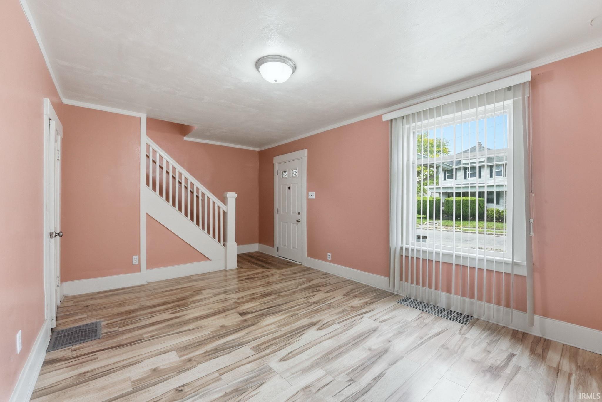 Spacious room featuring wood-finish flooring, a white staircase with balusters, a paneled entry door with decorative glass inserts, and a large window with vertical blinds