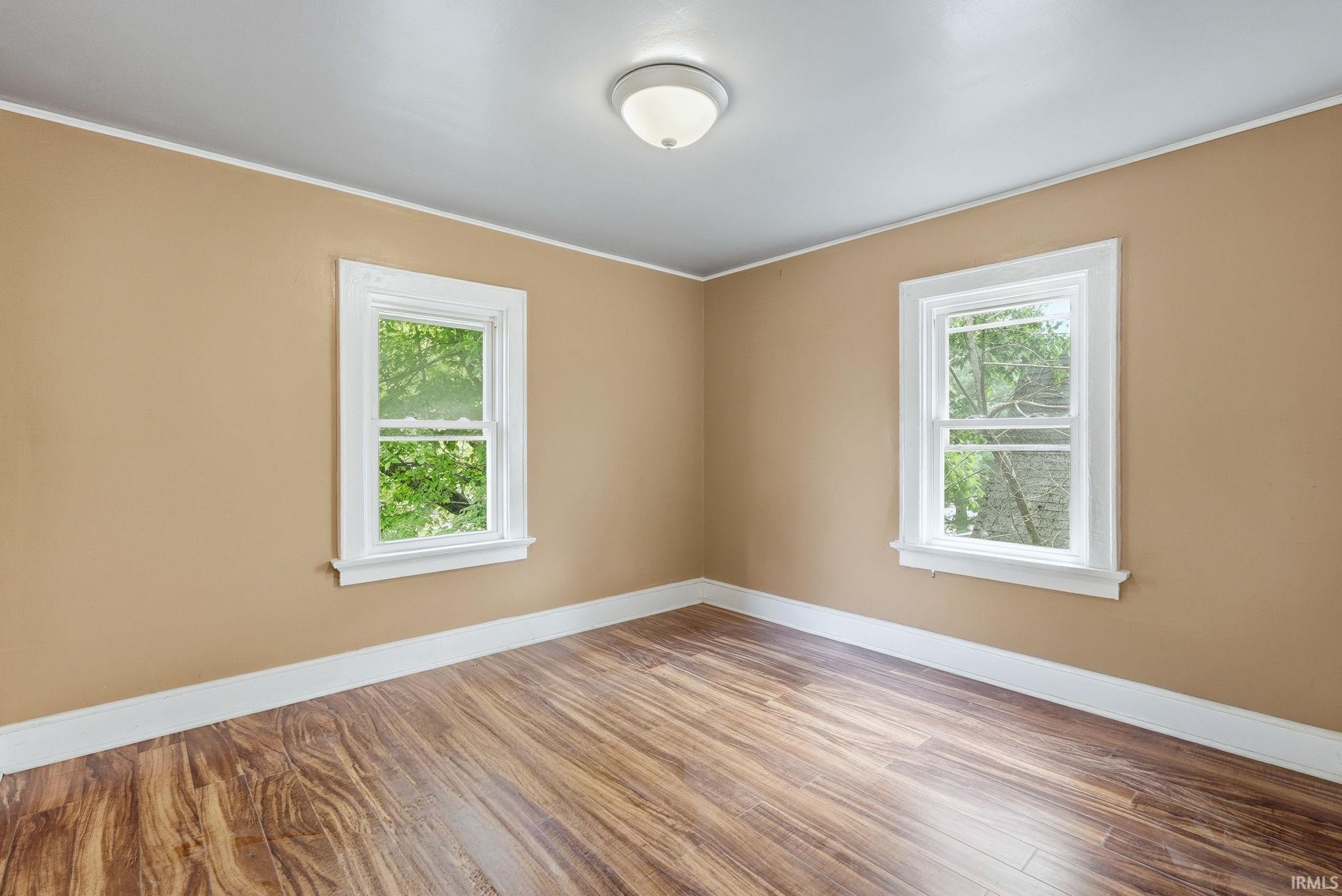 Room featuring two white-framed windows, wood-finish flooring, light tan walls, white trim, and a flush-mount ceiling light