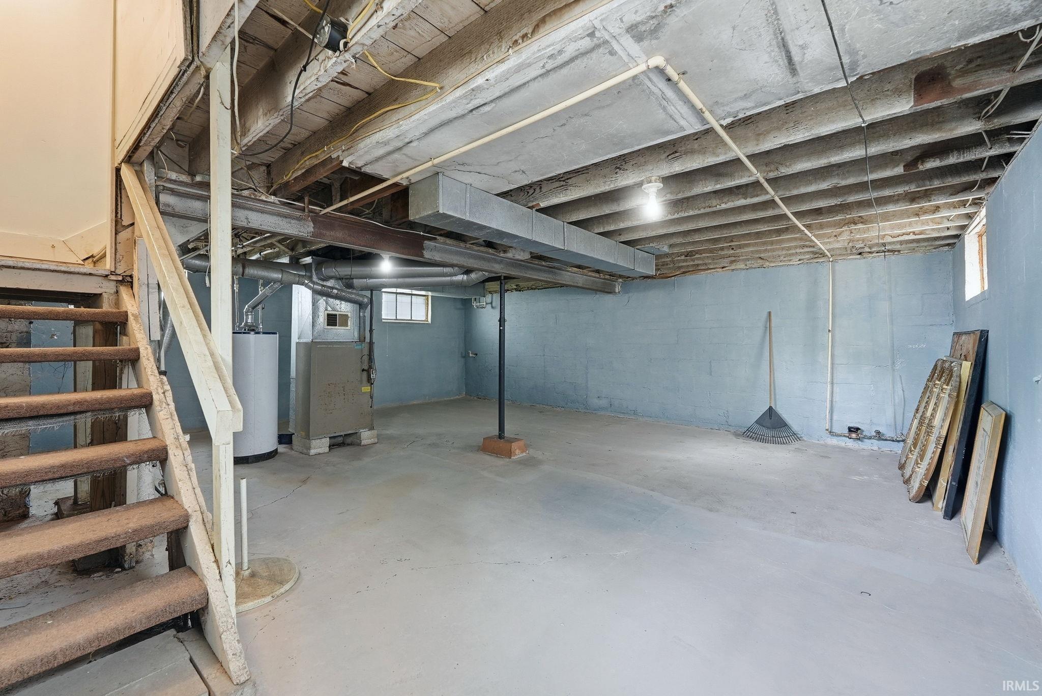 Unfinished basement featuring a concrete floor, painted cinder block walls, exposed wood ceiling joists, utility windows, and a water heater