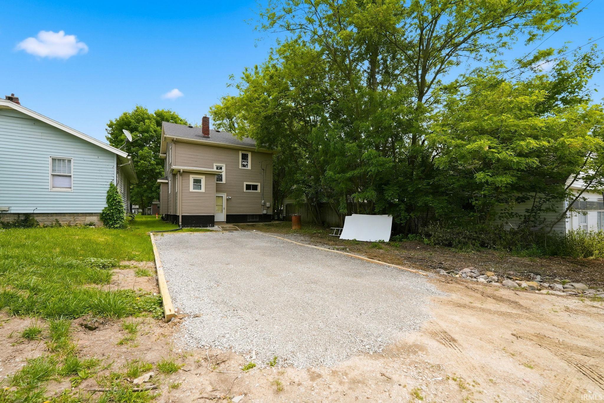 Spacious gravel parking area alongside the tan-sided property, featuring a white rear entry door and mature surrounding trees