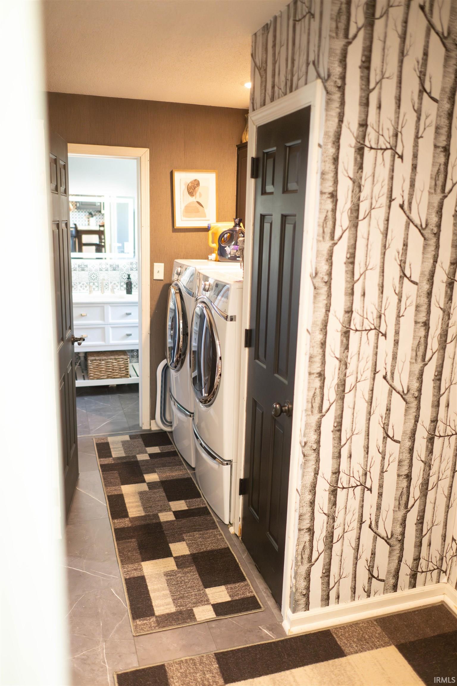 Dedicated laundry area featuring stacked front-loading appliances, grey tile flooring, and tree-patterned wall covering