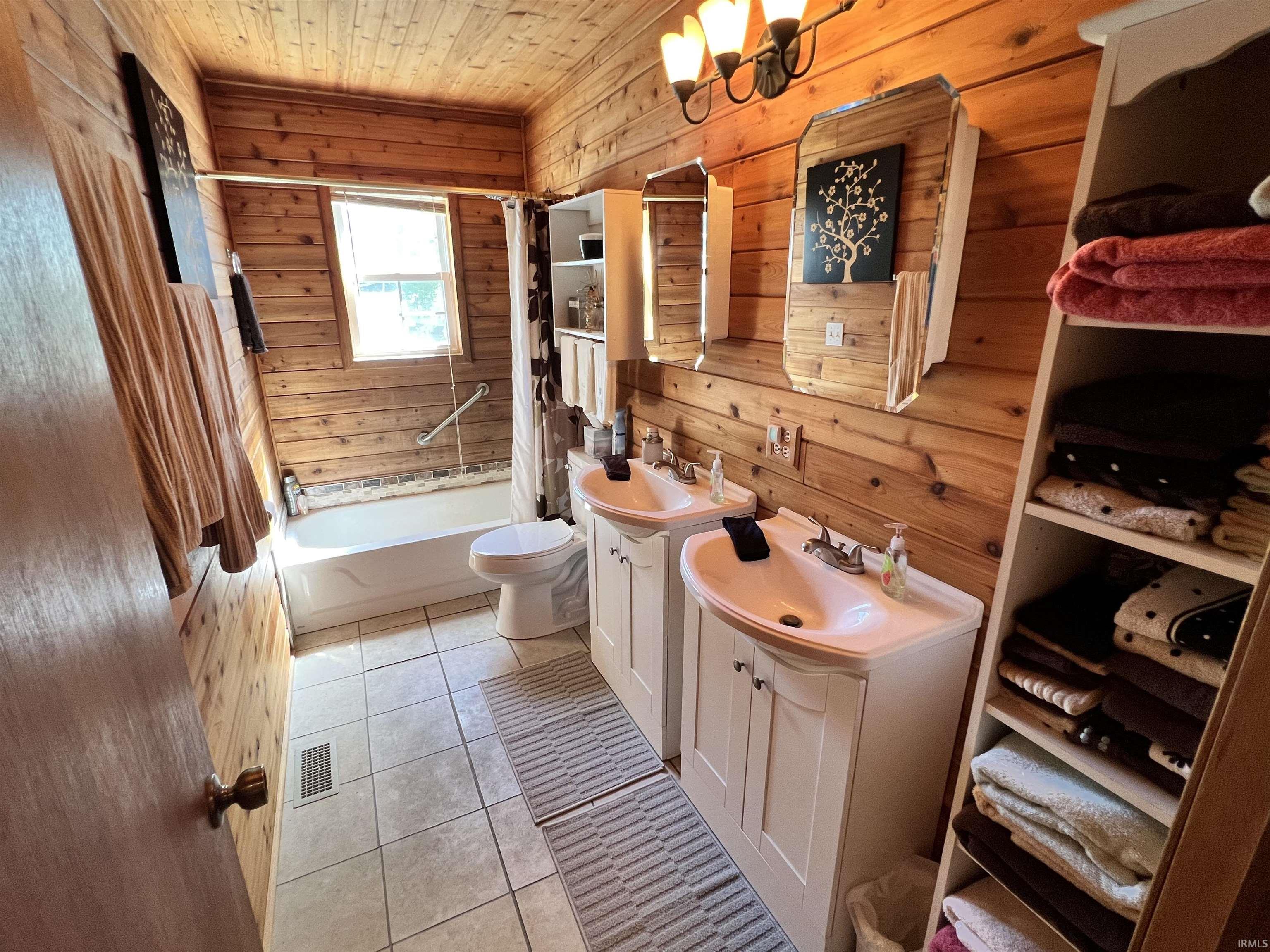 Bathroom featuring wood-paneled walls and ceiling, dual vanity sinks with white cabinetry, a built-in bathtub with shower, and tile flooring