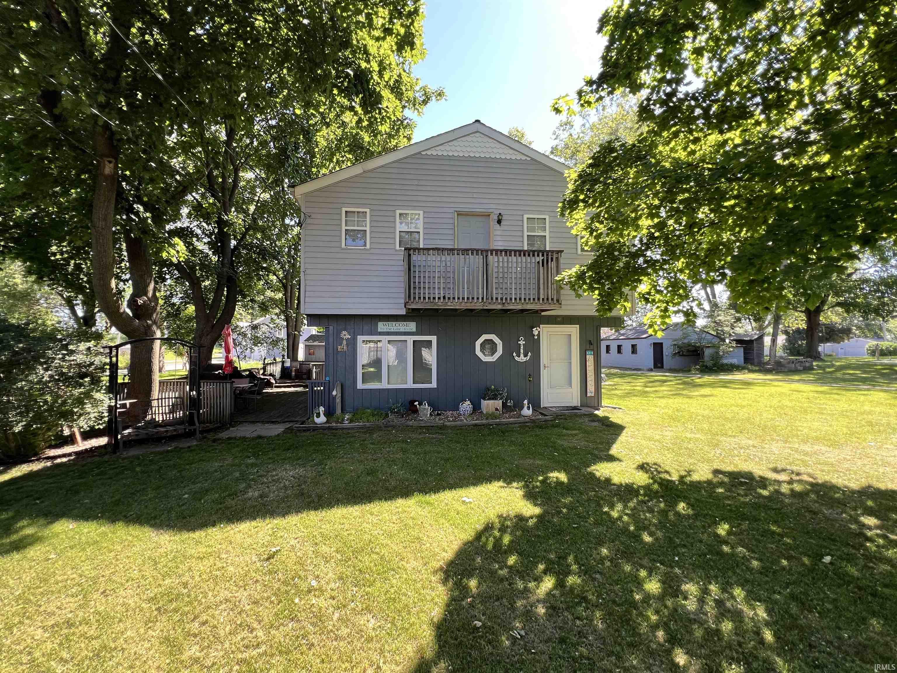 Two-story home featuring a full-width upper-level deck with wooden railings, a lower-level patio area, expansive lawn, and mature trees providing natural shade
