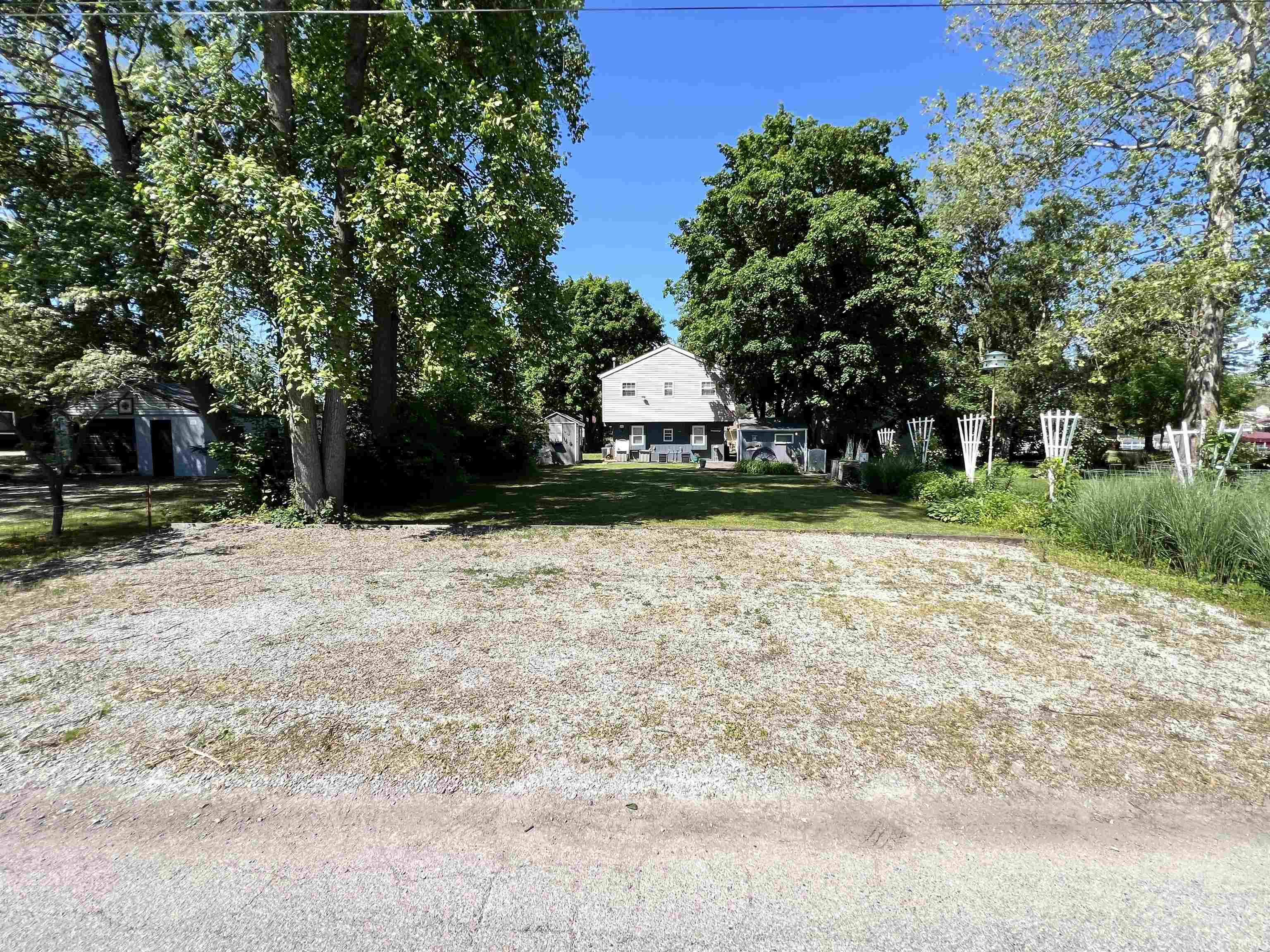 Gravel parking area fronting a property with a two-story residence, a detached shed, and mature trees