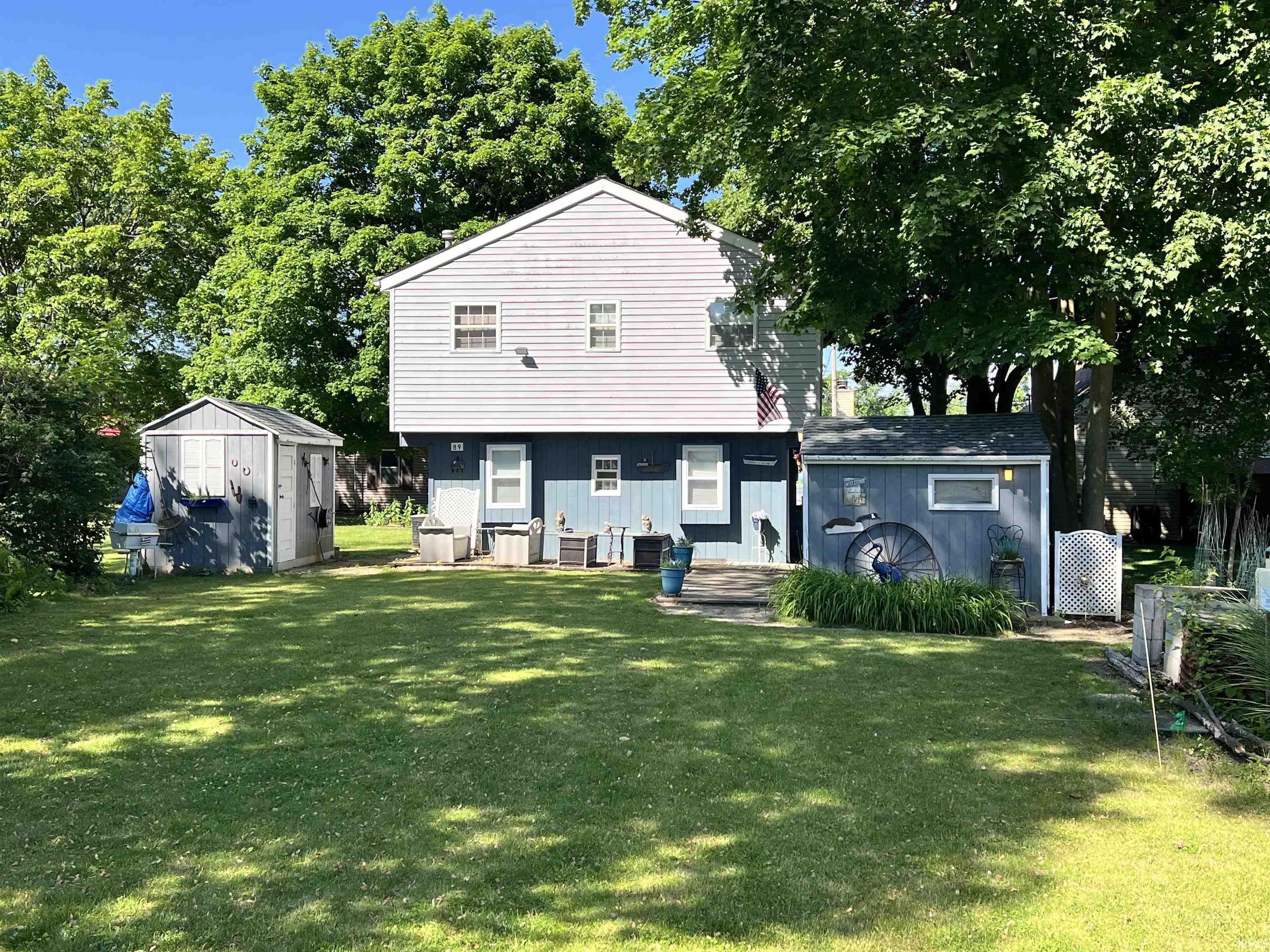 Spacious backyard featuring a manicured lawn, two detached storage sheds, mature perimeter trees, and a ground-level patio area