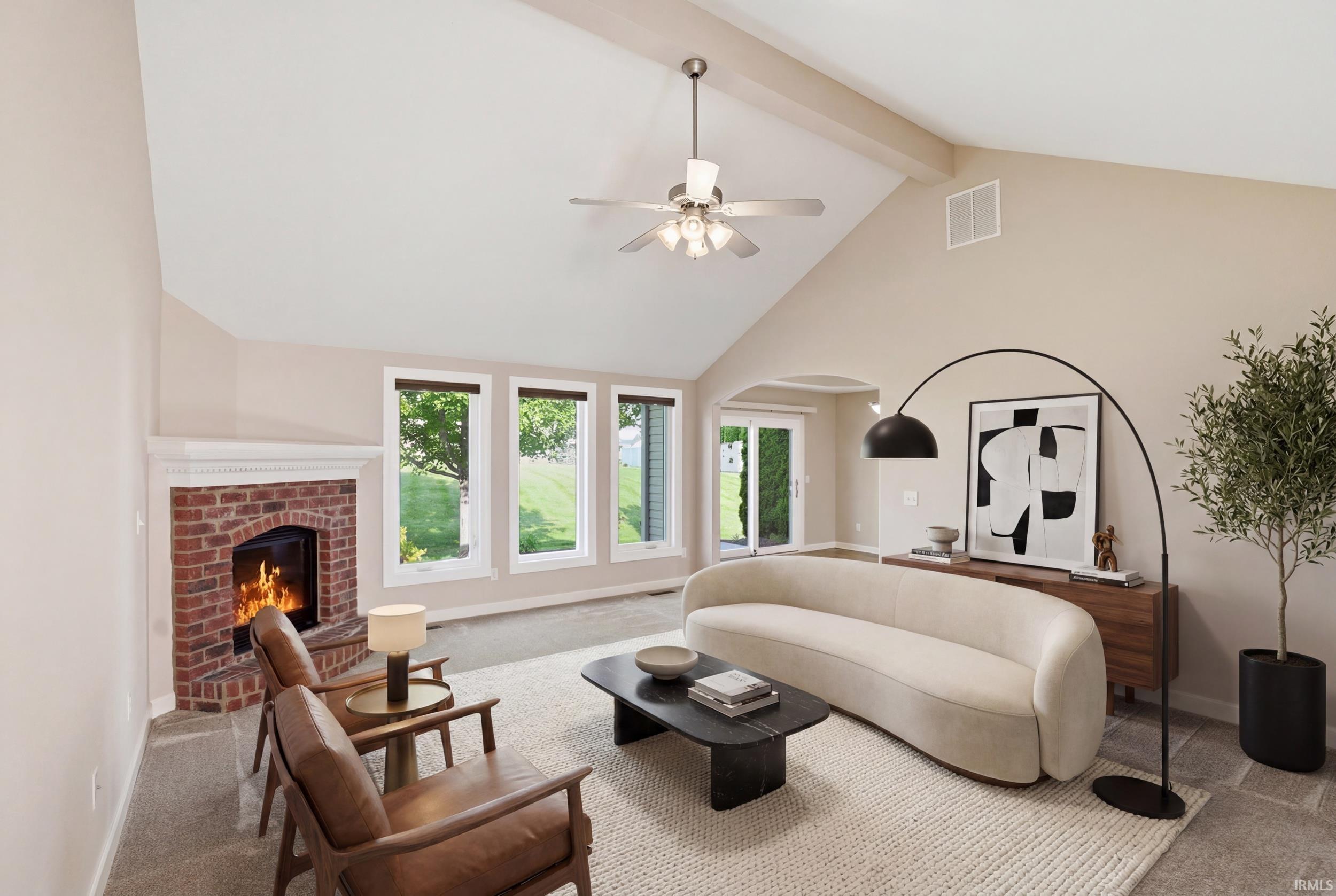 Spacious living area featuring a vaulted ceiling with exposed beam, a ceiling fan, a brick fireplace with a white mantel, and multiple windows