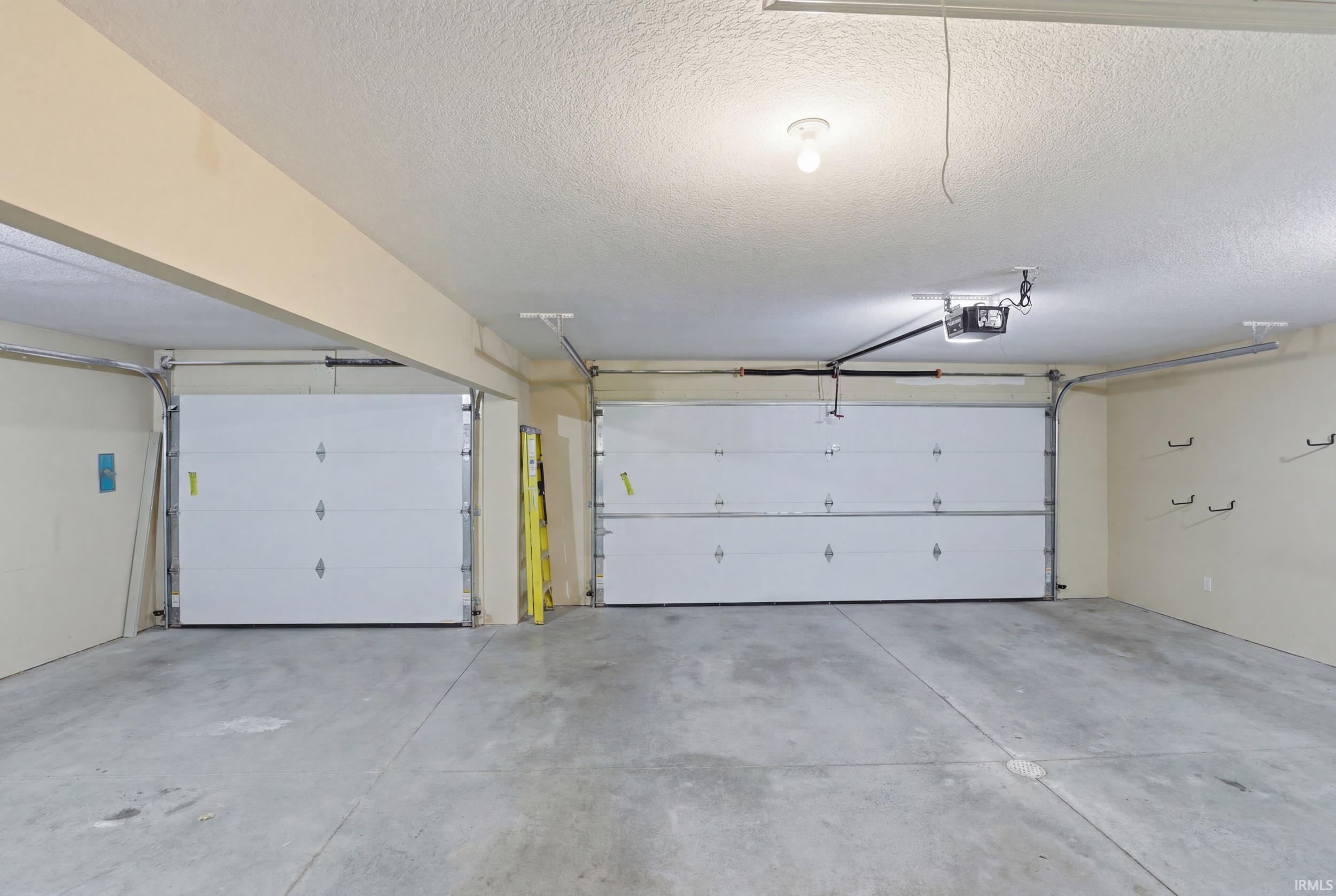 Two white paneled garage doors, concrete flooring, overhead lighting, and wall-mounted hooks for storage