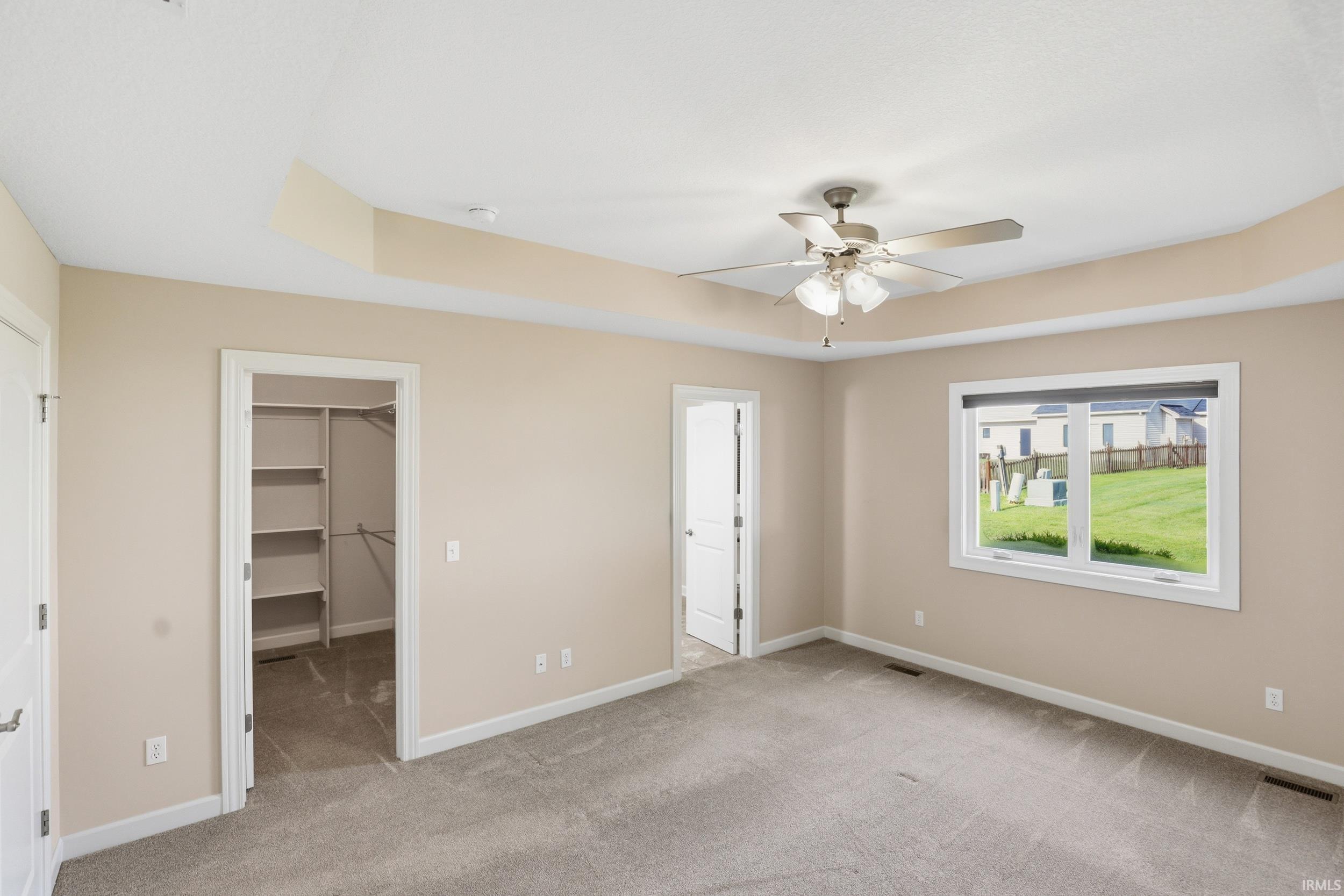 Spacious room featuring a tray ceiling, ceiling fan with light fixture, light-colored carpeting, a walk-in closet with built-in shelving, and a large window