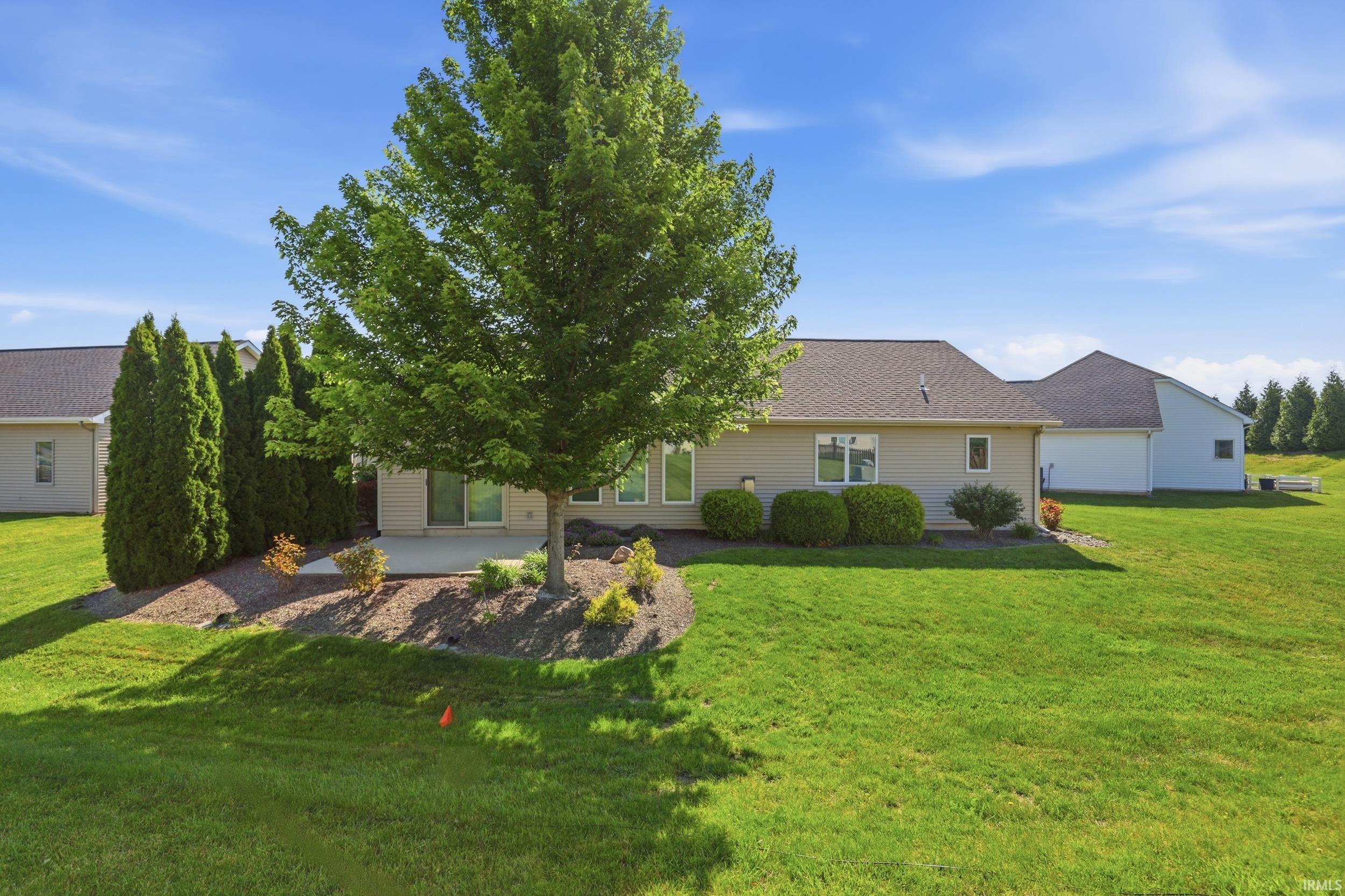 Rear exterior featuring a concrete patio with landscaping, mature trees, and a spacious lawn