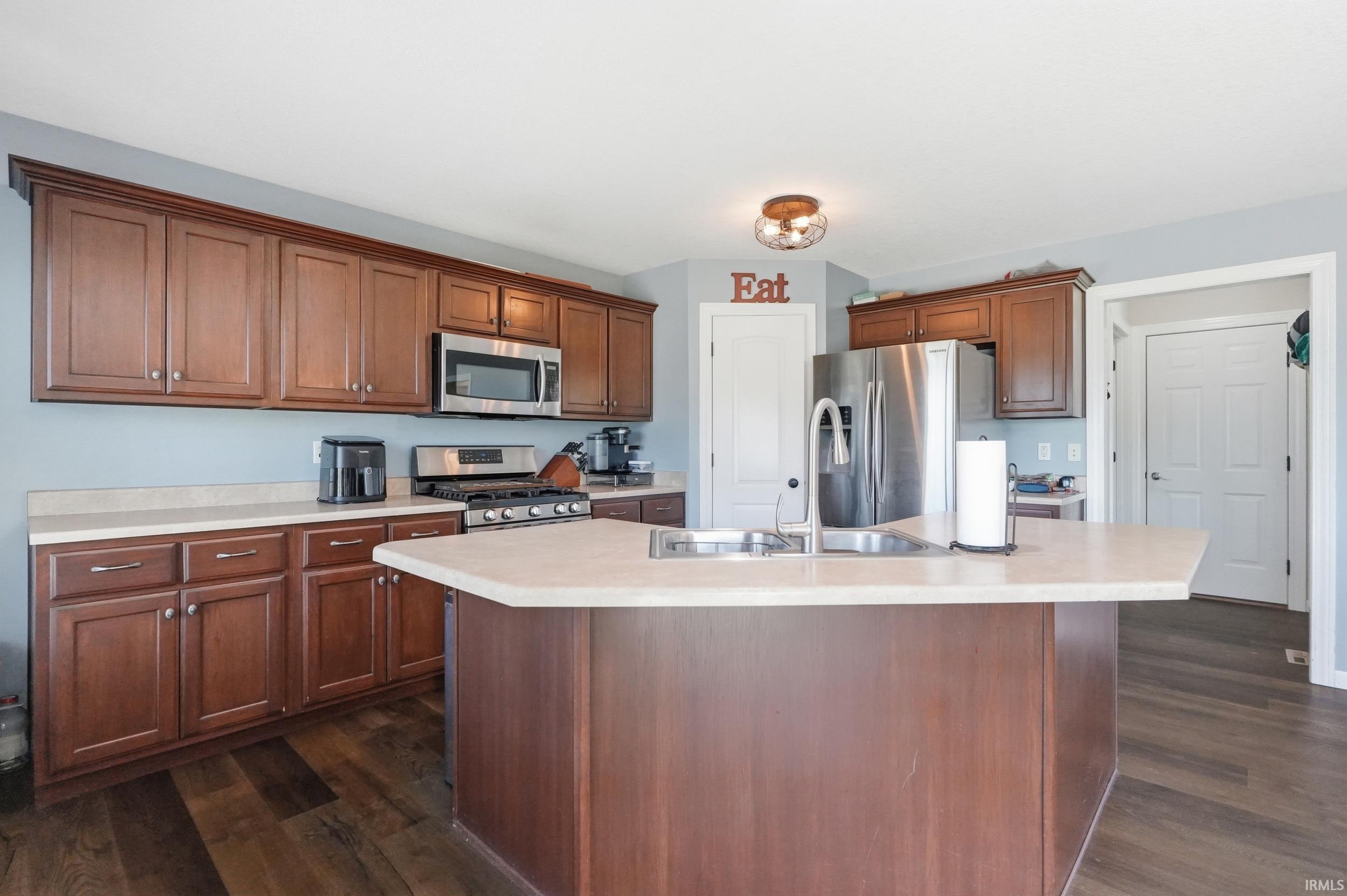 Kitchen featuring a central island with an undermount sink, rich wood cabinetry, stainless steel appliances, light countertops, and wood-finish flooring