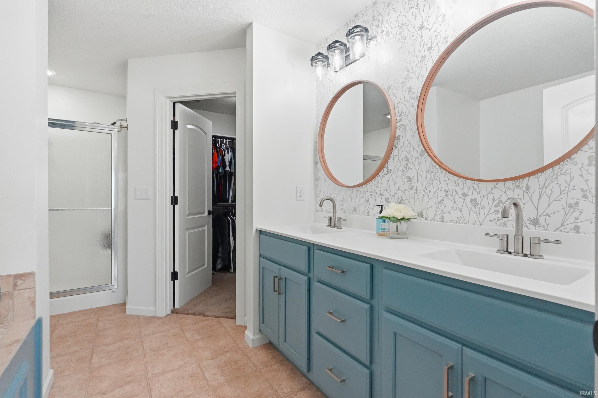 Bathroom featuring a dual vanity with a white countertop and blue cabinetry, round mirrors, textured wallpaper, and a glass-enclosed shower