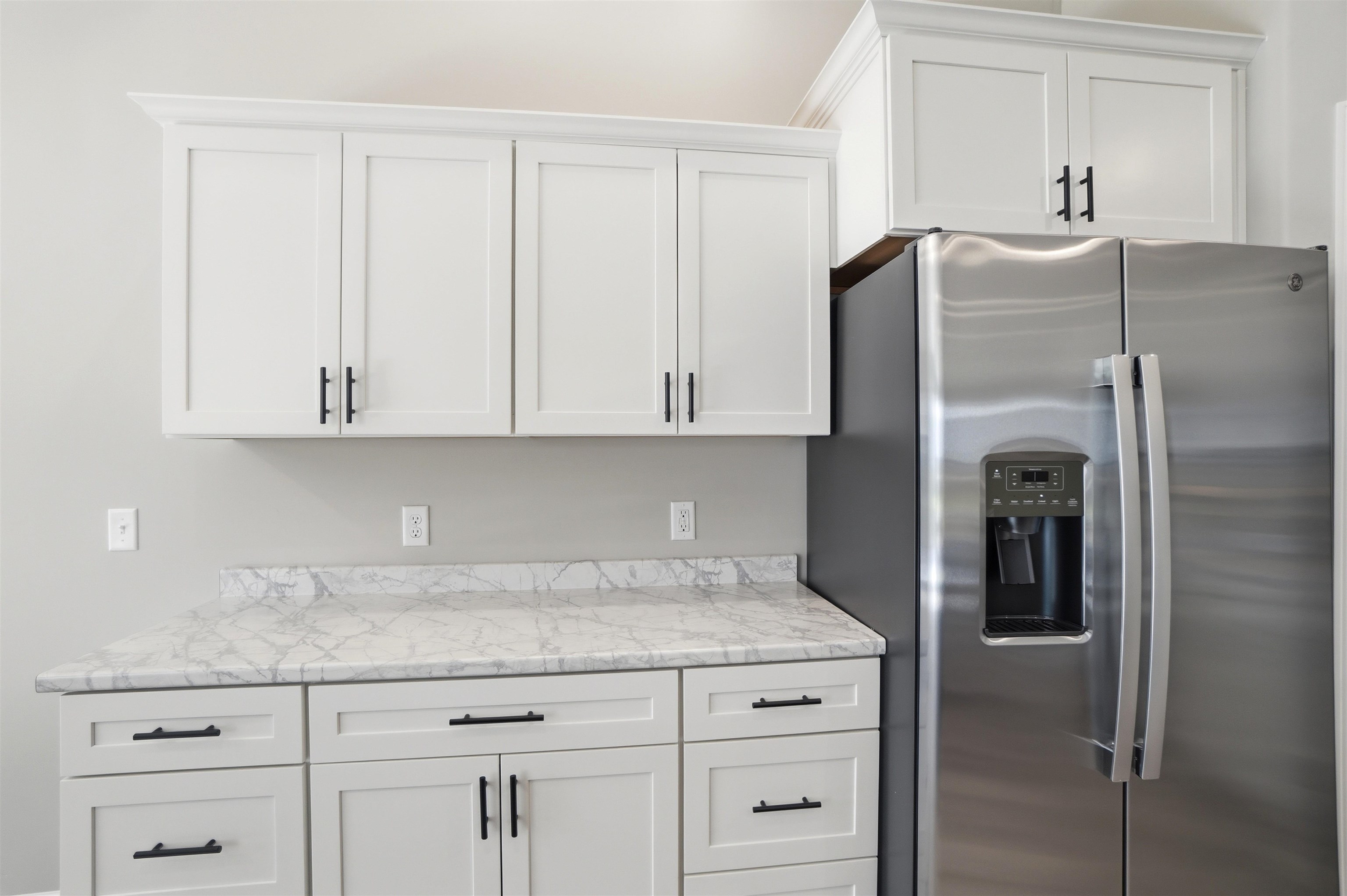 White shaker-style cabinetry with matte black hardware, stone-finish countertops, stainless steel refrigerator, and crown molding accents