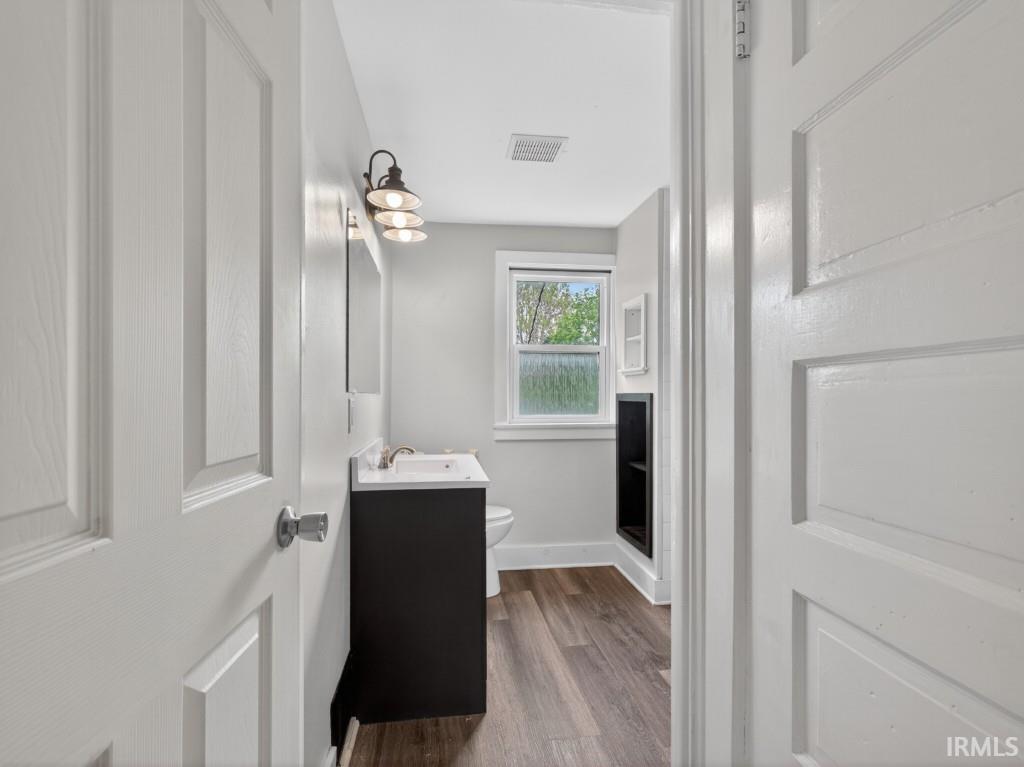 Bathroom featuring a dark wood-finish vanity with a white countertop and basin sink, industrial-style wall sconce, rectangular mirror, wood-finish flooring, and a window with privacy glass