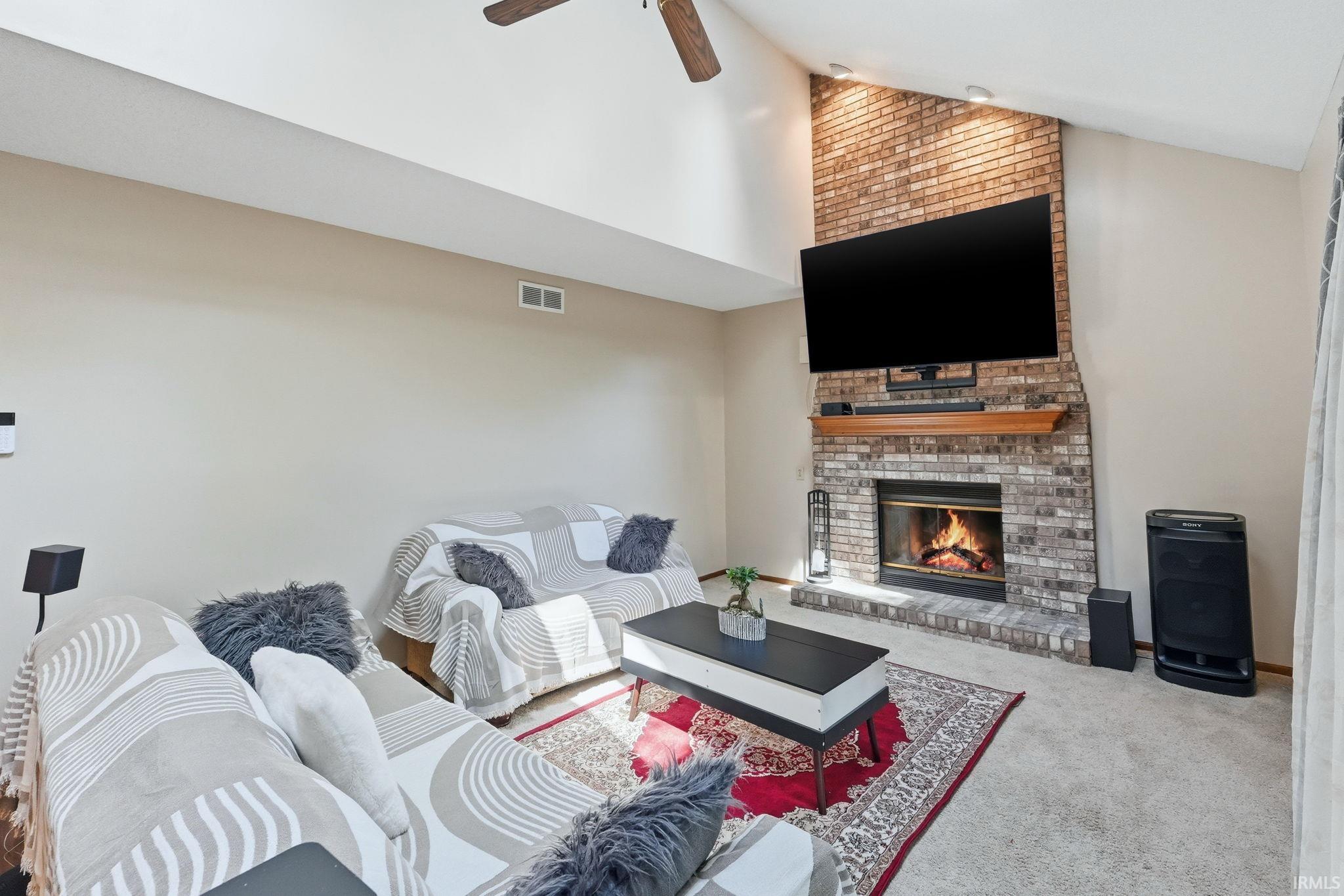 Living area featuring a floor-to-ceiling brick fireplace with a wood mantle, vaulted ceiling with a ceiling fan, and light-toned wall finishes