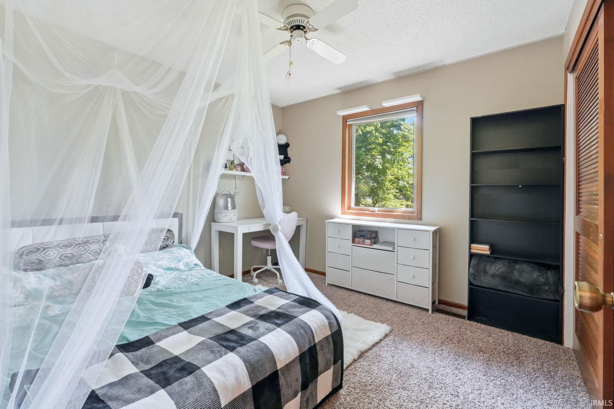 Carpeted bedroom featuring a ceiling fan, a window with wood trim, built-in shelving, and a louvered bifold closet door