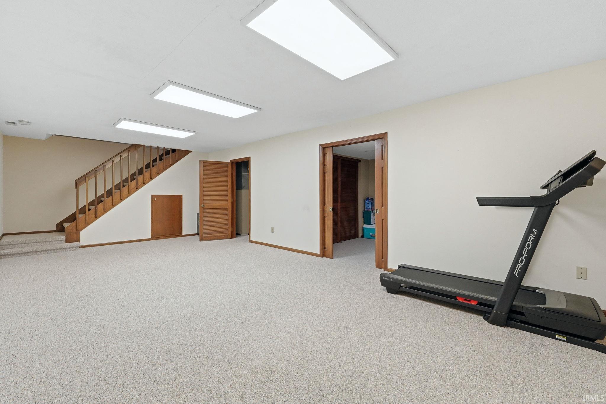 Spacious finished room featuring light-colored carpeting, multiple rectangular ceiling lights, and a prominent wood staircase with matching railing