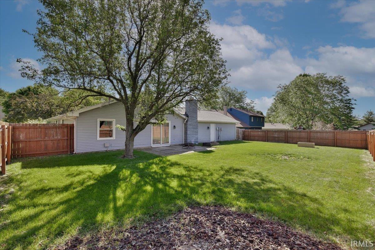 Generous, fenced backyard featuring a large tree, manicured lawn, and a light-colored patio