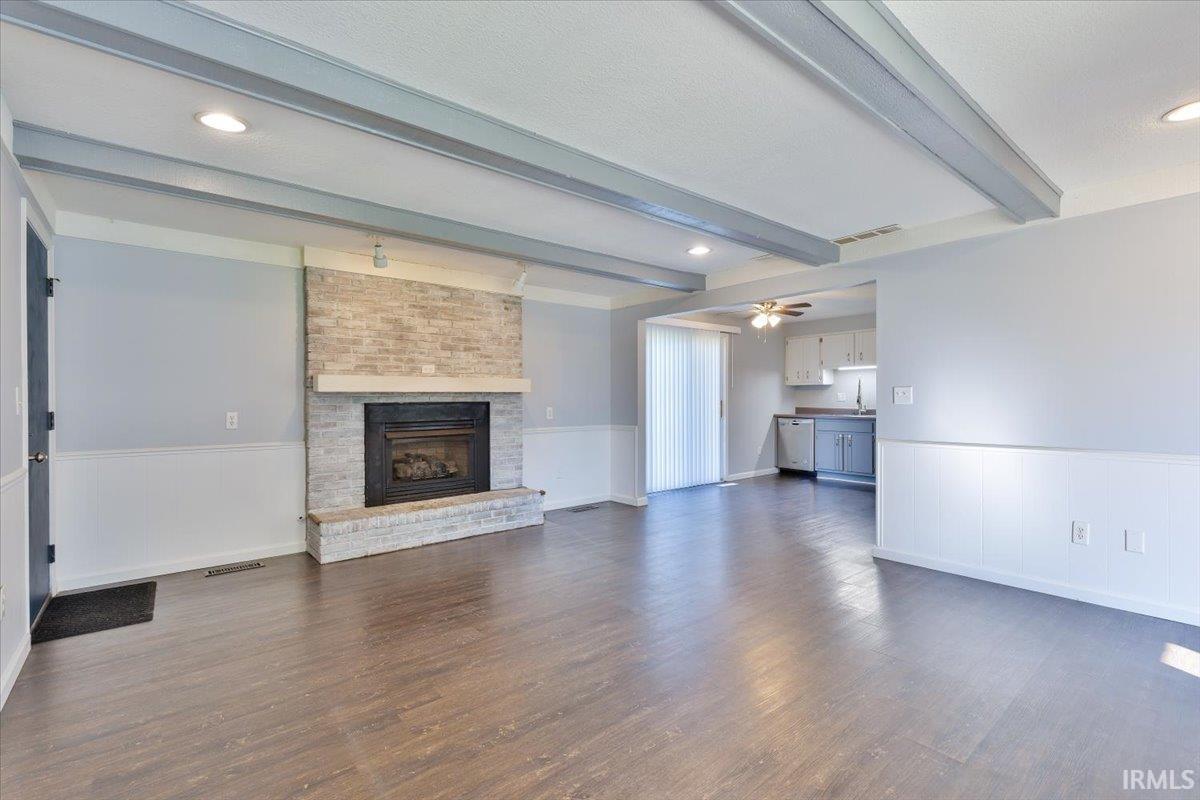 Spacious living area featuring a stone fireplace with a substantial mantel, wood-finish flooring, and exposed ceiling beams