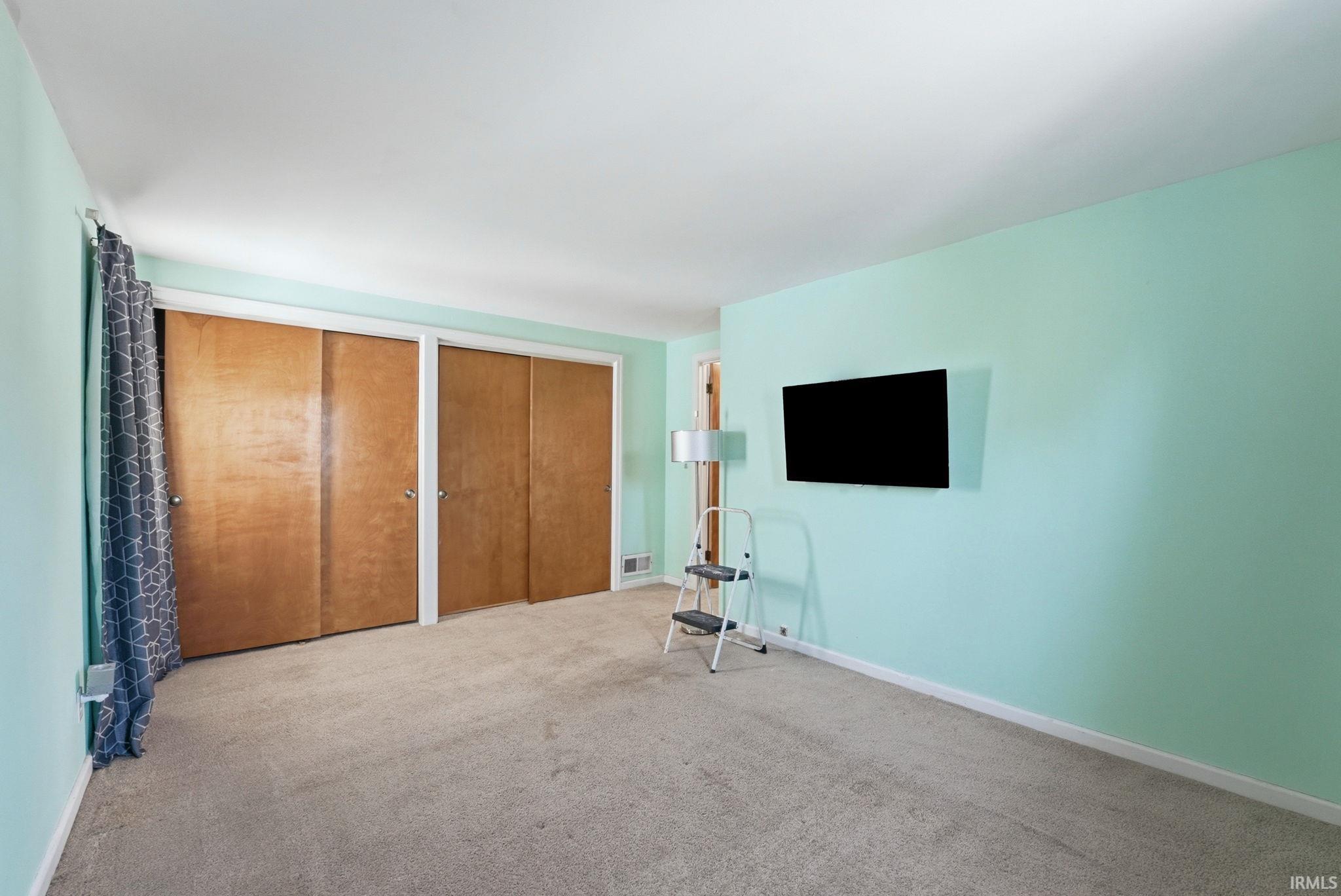 Room featuring light-colored carpeting, a wall-mounted television, and multiple closet doors with wooden finishes