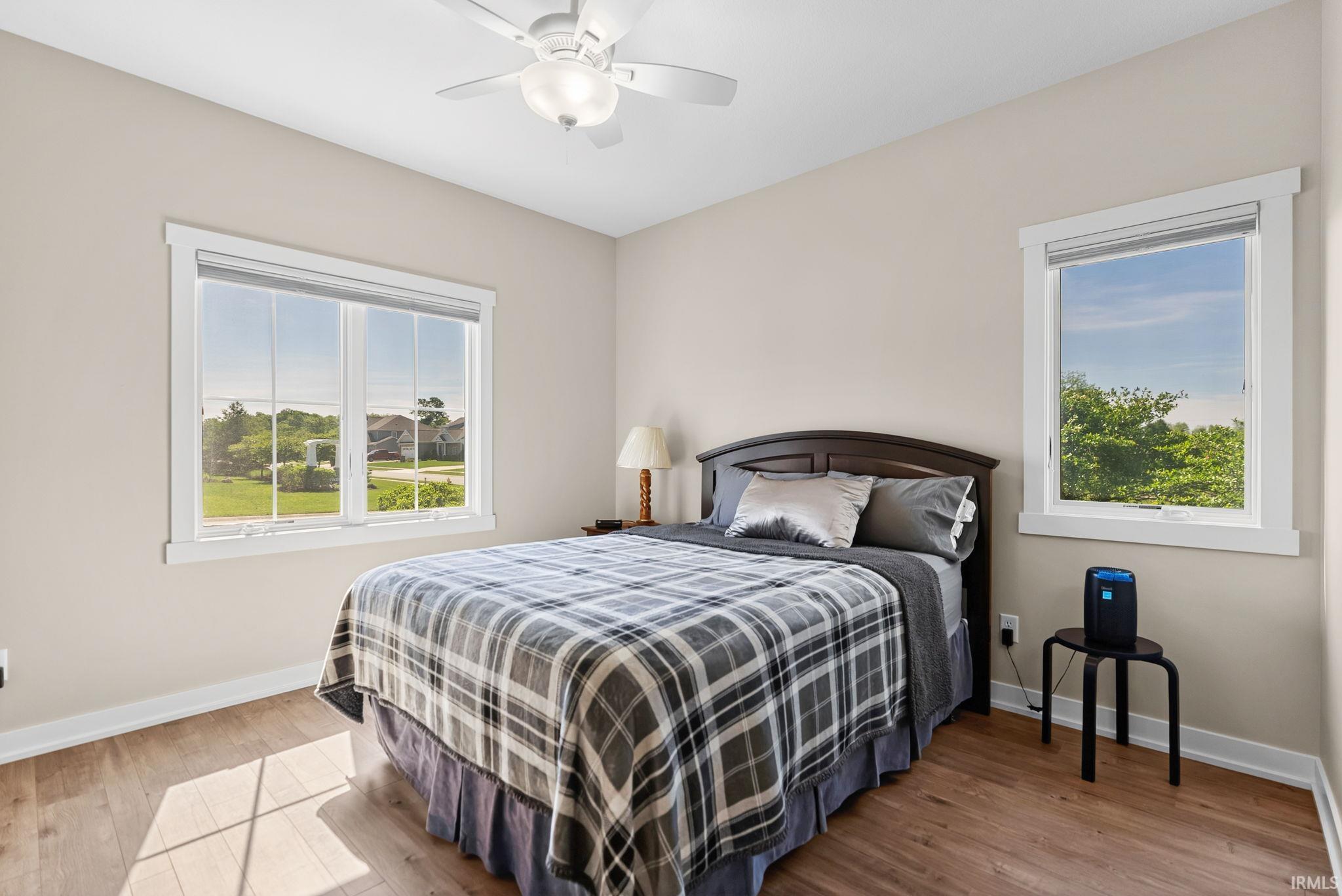 Bedroom featuring wood-finish flooring, two windows with white trim, light-toned wall paint, and a ceiling fan with integrated lighting