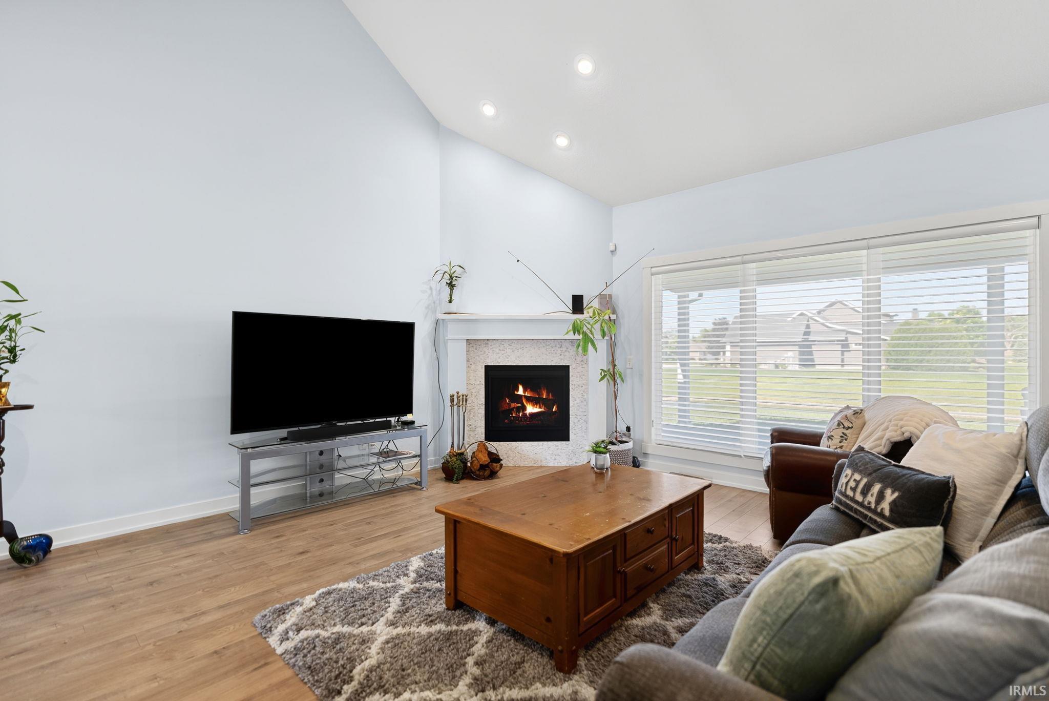 Vaulted ceiling living room featuring a fireplace with a light-colored surround, wood-finish flooring, and expansive windows