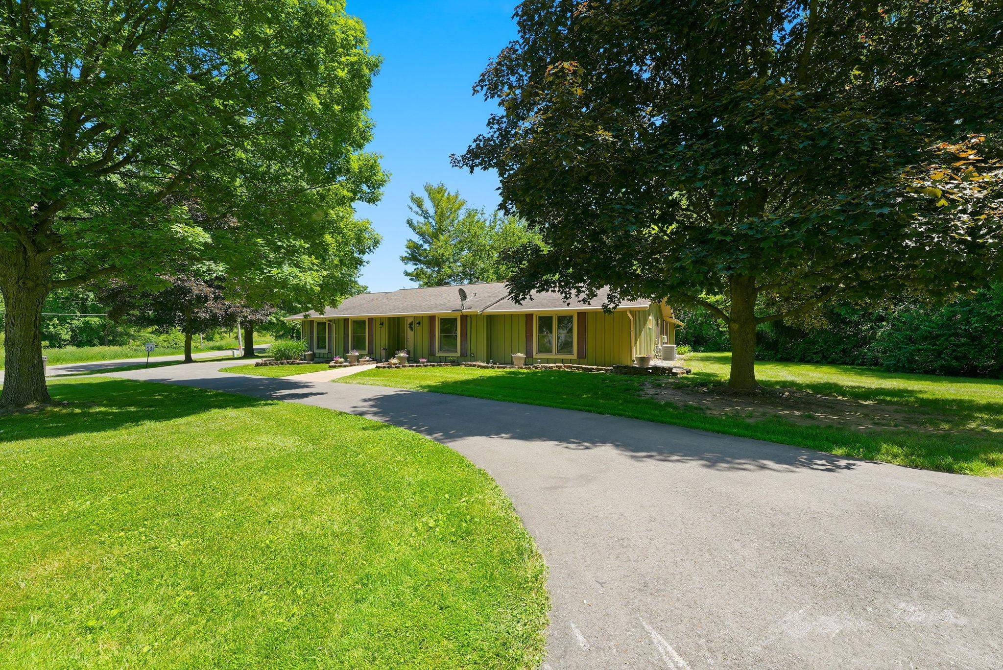 Ranch-style residence featuring a light green exterior, a paved circular driveway, and mature shade trees