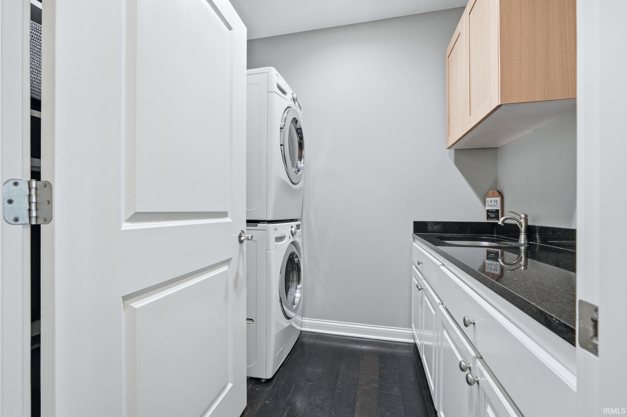 Dedicated laundry area featuring stacked front-load machines, a dark wood-finish floor, white base cabinetry with brushed nickel hardware, a dark countertop with an undermount sink, and light wood-tone upper cabinetry