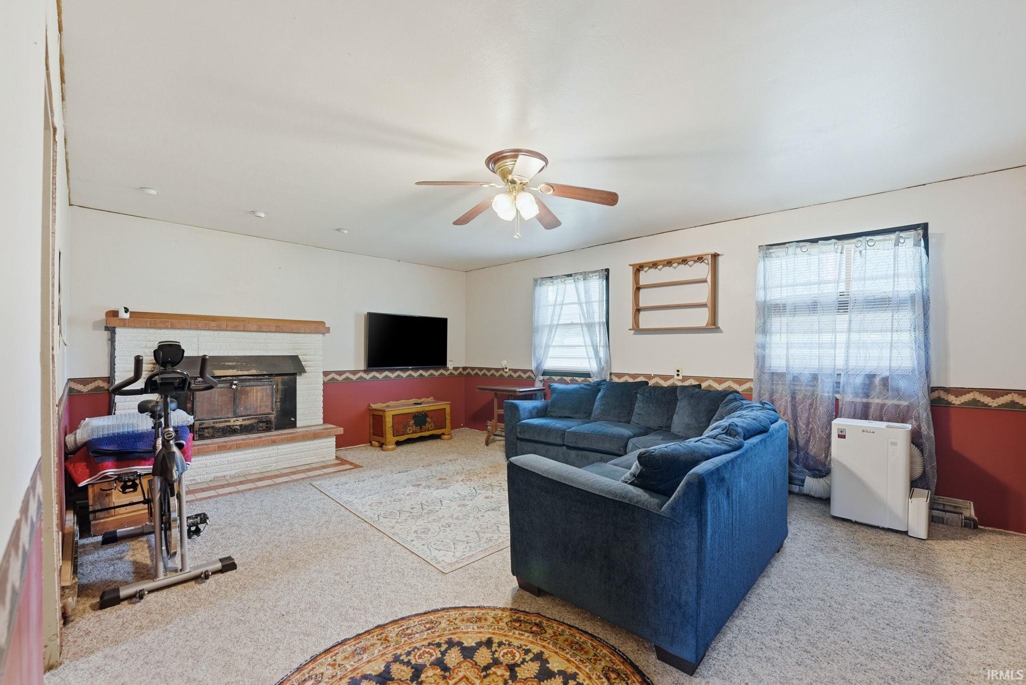 Living area featuring a brick hearth fireplace with a wood mantle, carpet flooring, a ceiling fan, and multiple windows