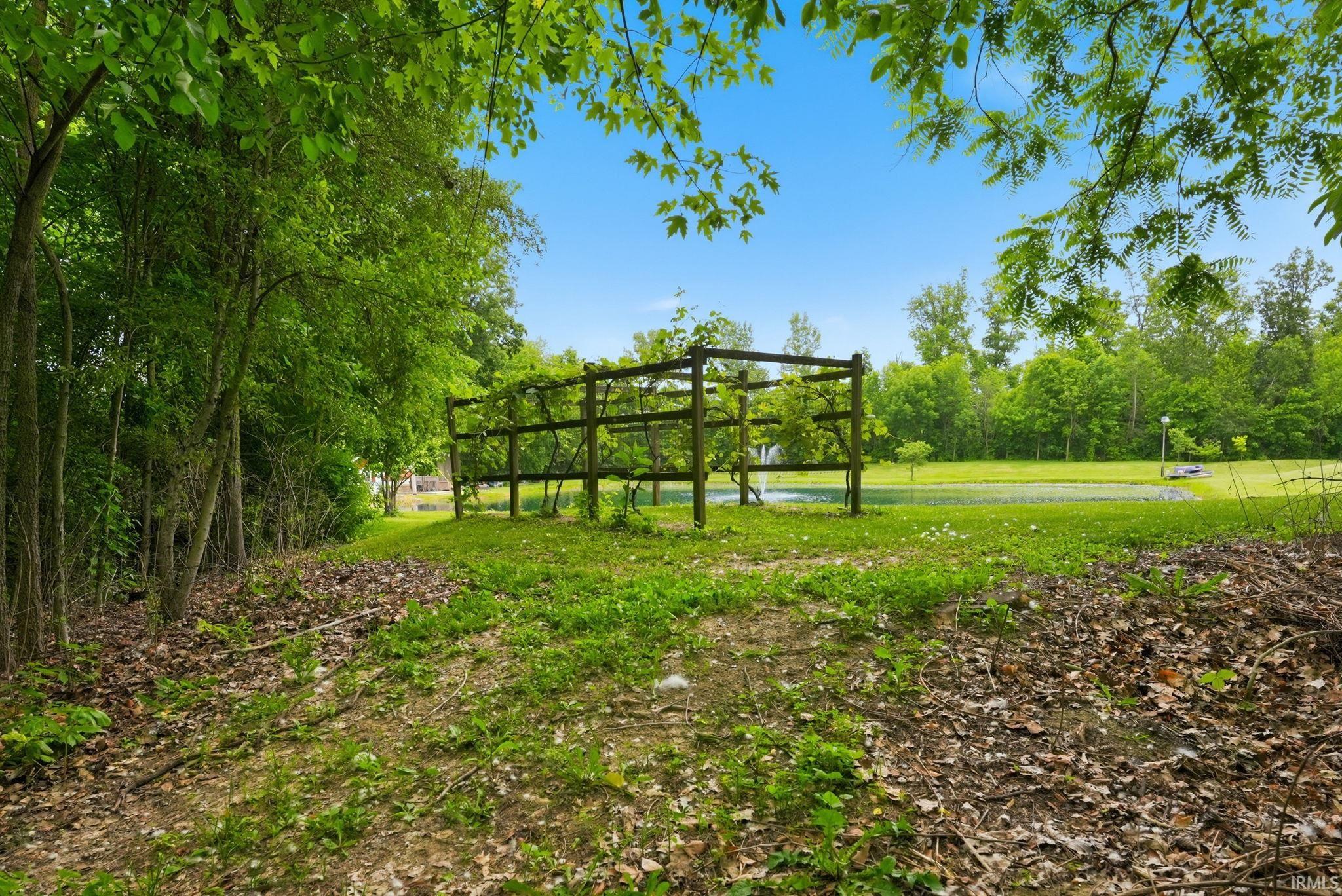 Expansive green space featuring a wooden arbor structure, mature trees, a natural pond, and a clear blue sky