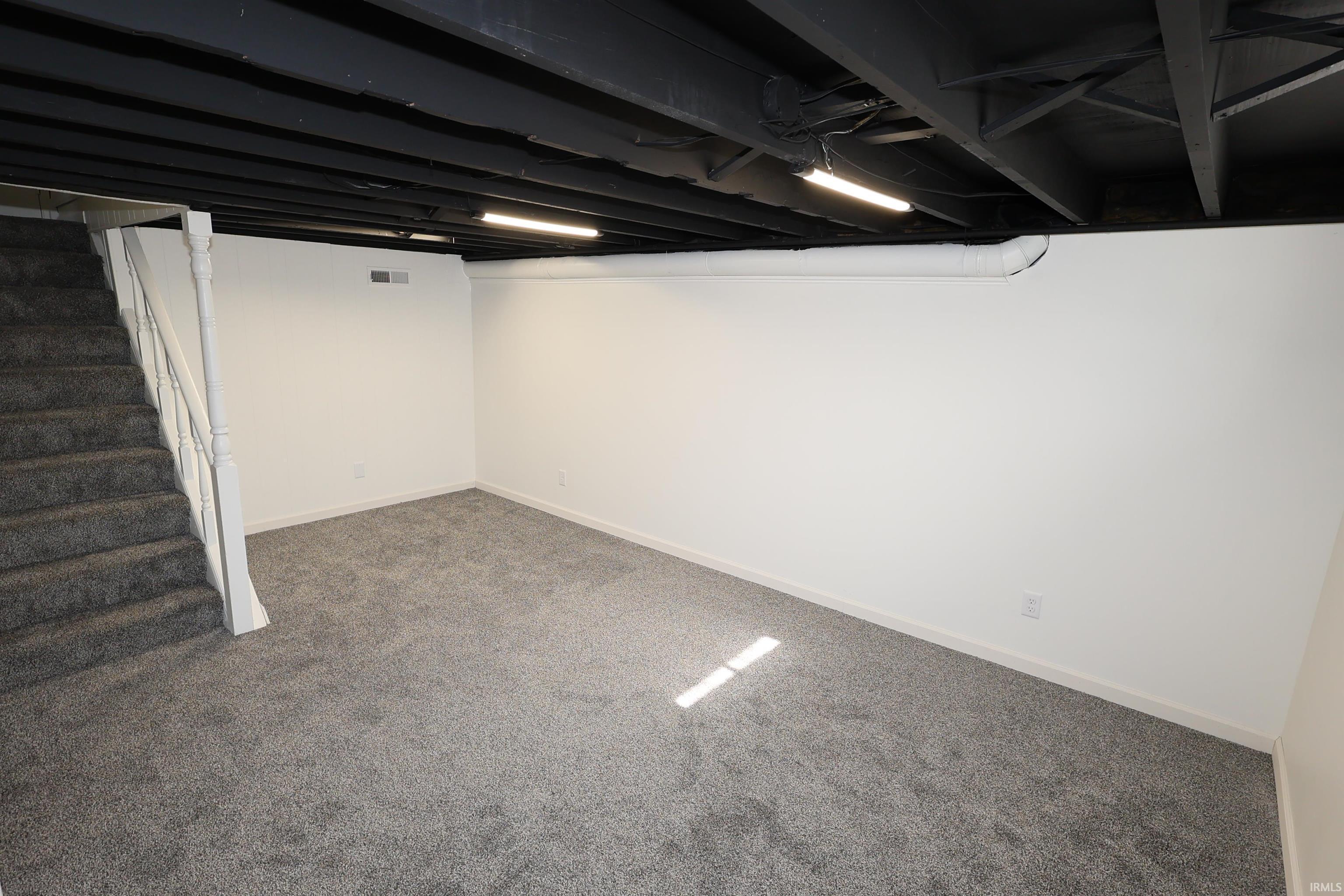Spacious lower-level room featuring new grey carpeting, crisp white walls, and a painted black exposed ceiling