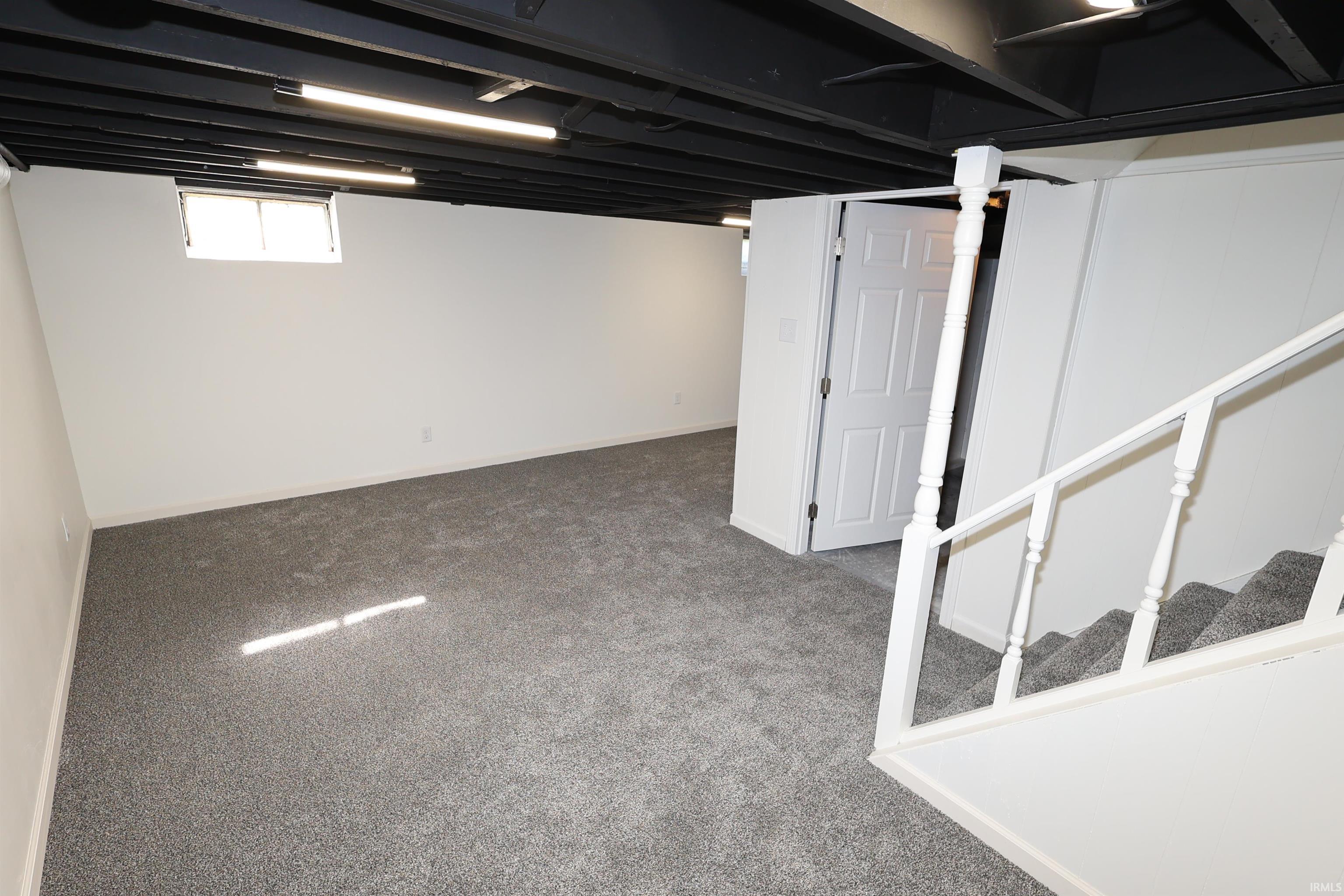 Finished lower level featuring new gray carpet, white walls, painted black exposed ceiling joists with linear light fixtures, and a white staircase with balusters