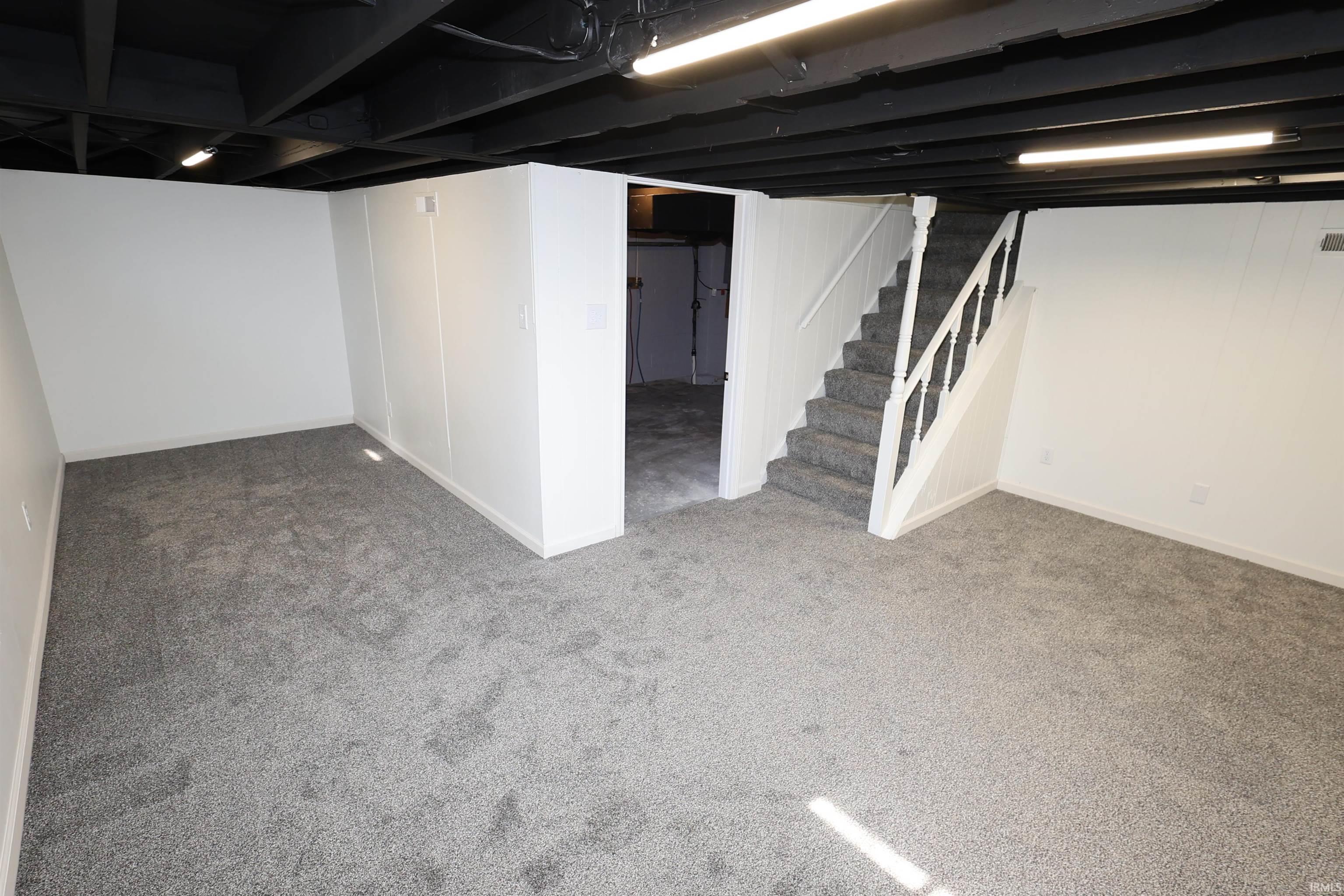 Spacious lower-level area with grey carpet flooring, white walls, and a contrasting black exposed ceiling with strip lighting