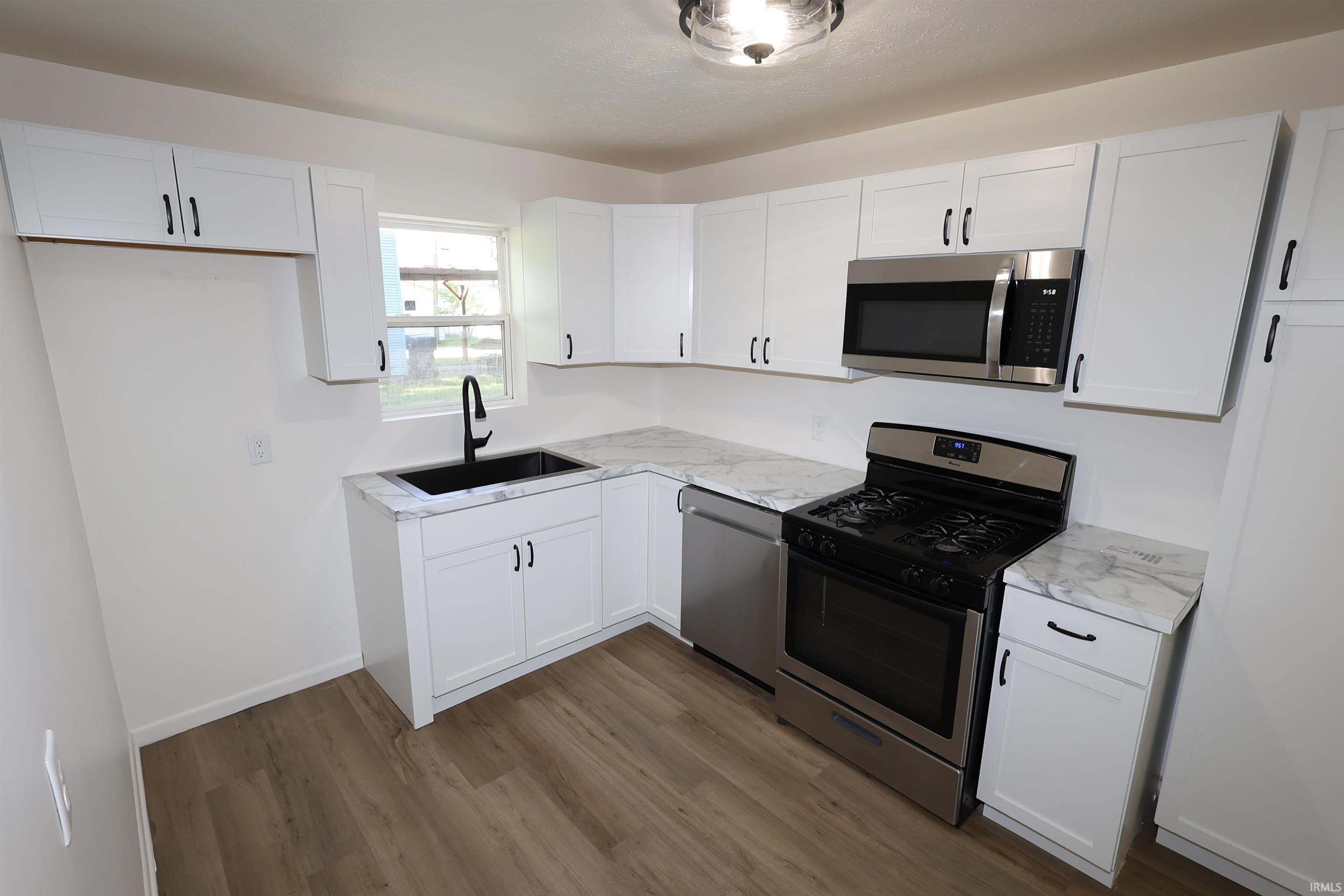 Kitchen featuring white shaker-style cabinetry, wood-finish flooring, stainless steel appliances, a black basin sink with a matte black faucet, and stone-finish countertops