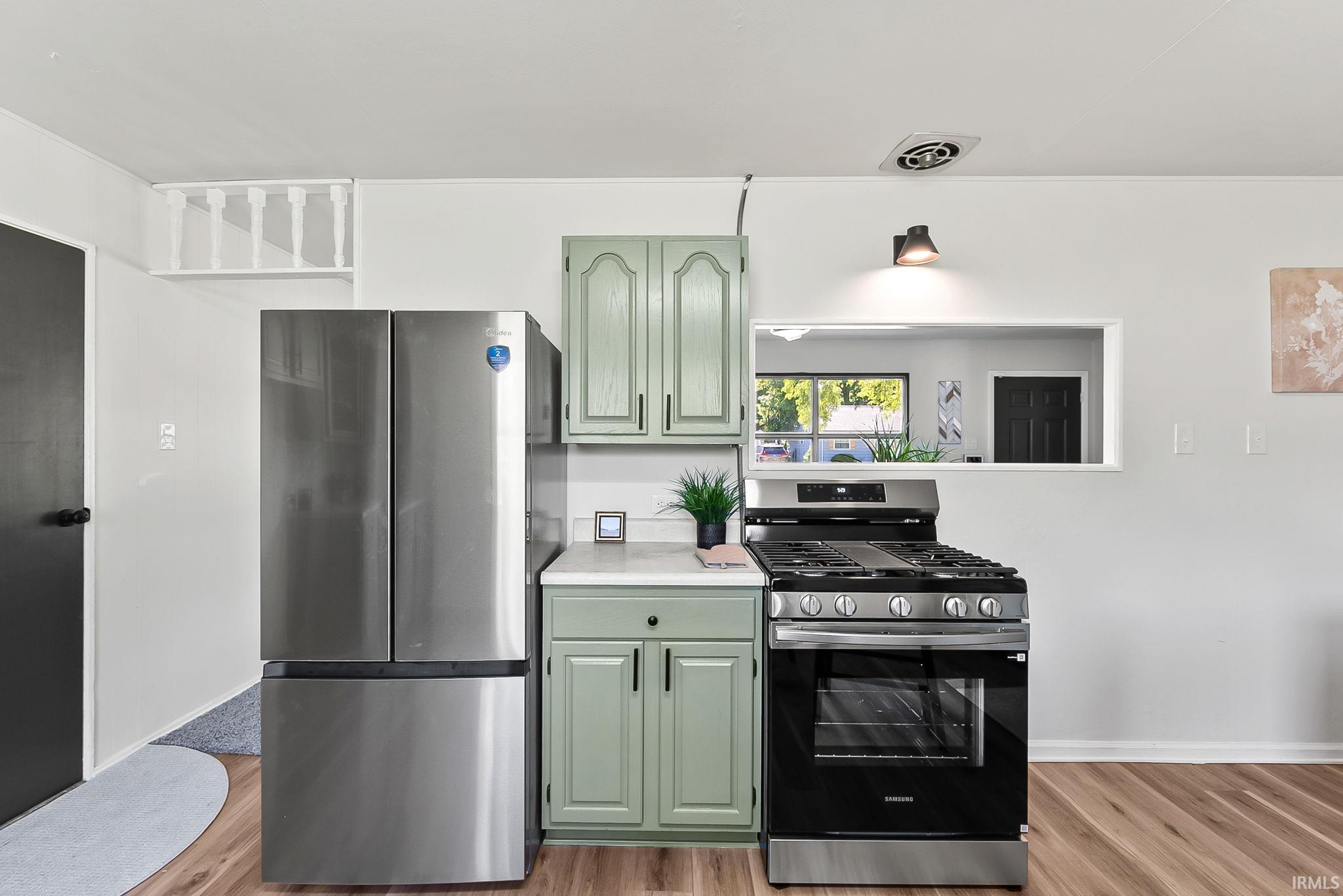 Modern kitchen featuring wood-finish flooring, light green cabinetry with matte black hardware, stainless steel appliances, and a pass-through window