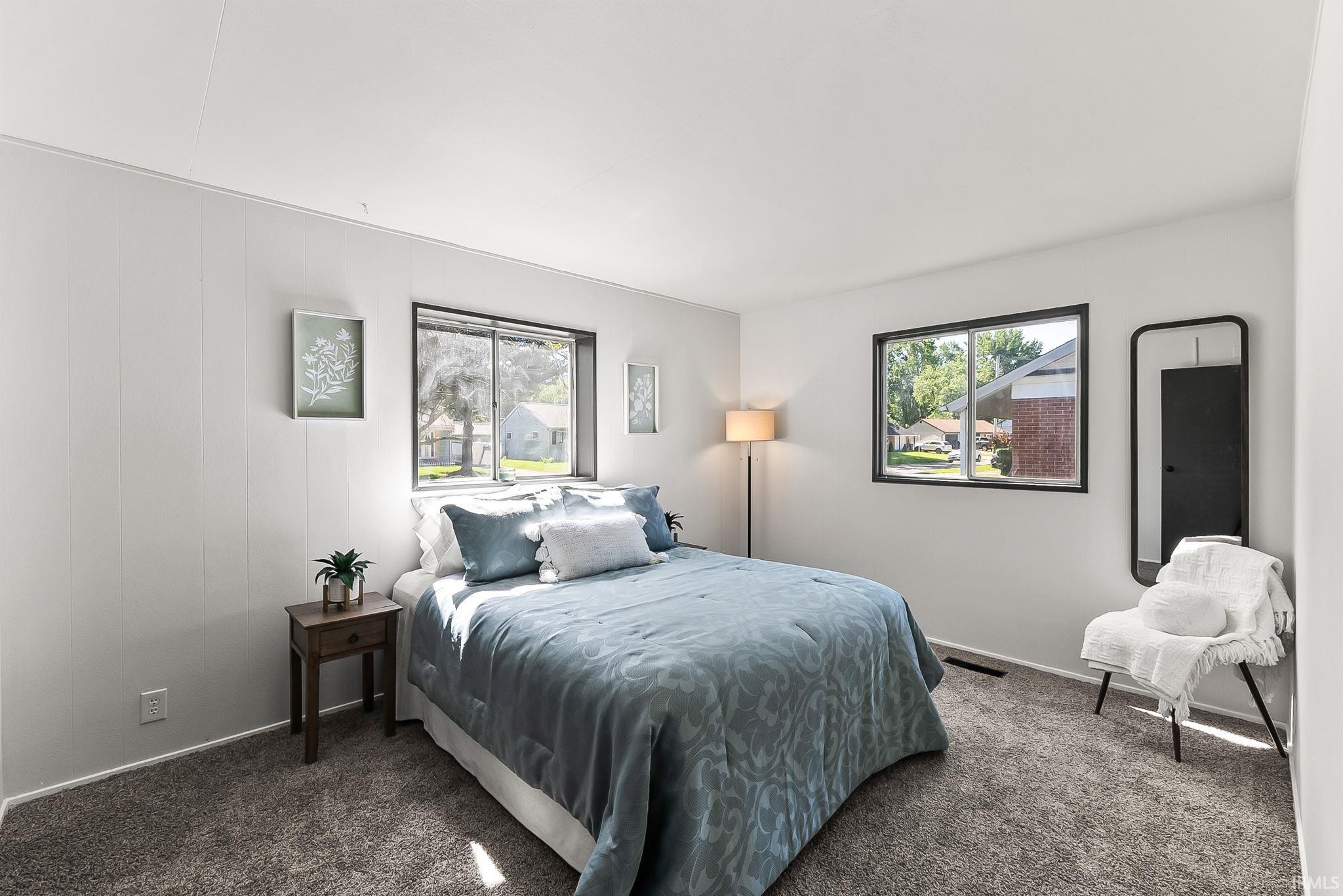 Spacious bedroom with two windows, showcasing light gray paneled walls and plush gray carpeting