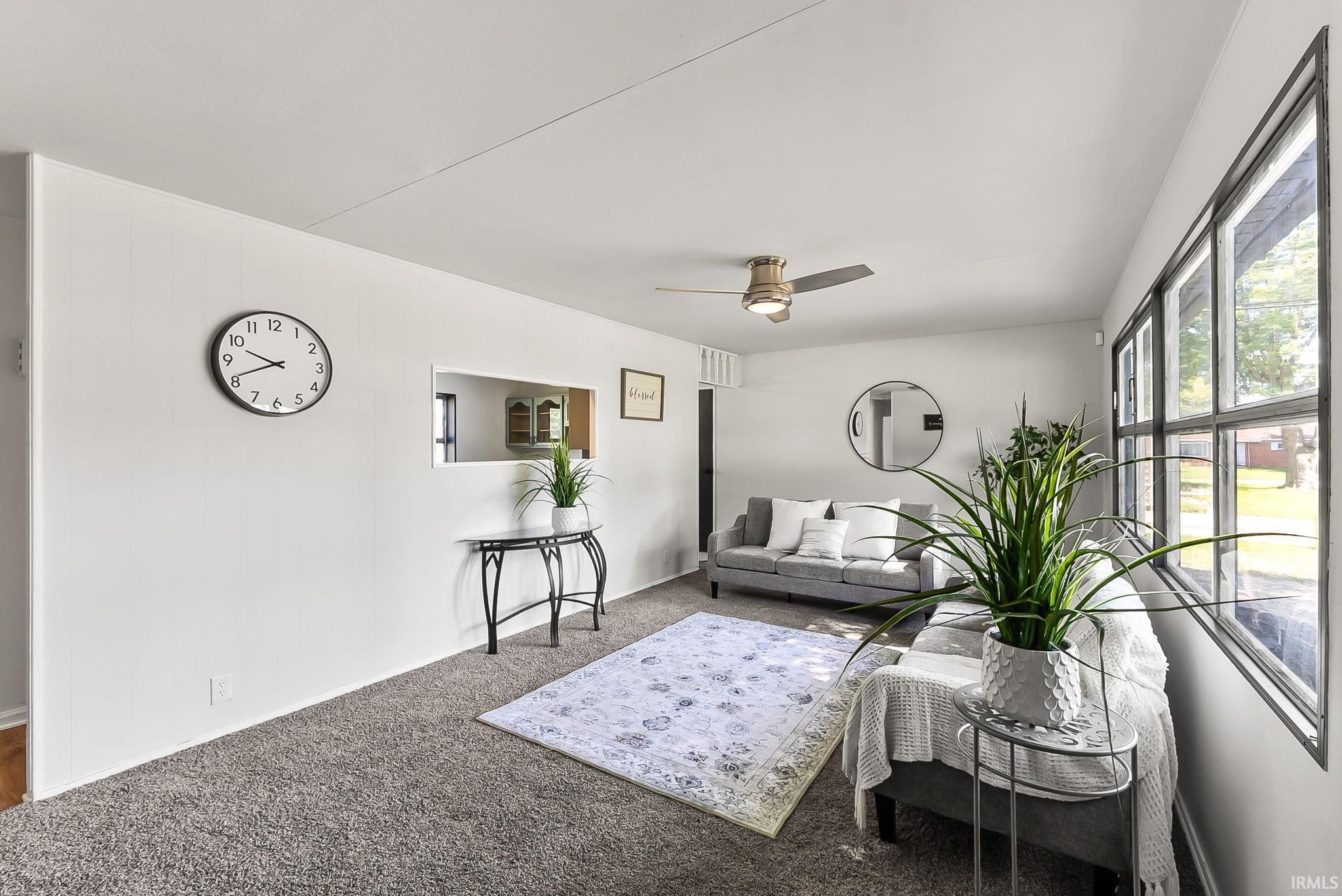 Spacious living area featuring neutral-toned carpeting, ample natural light through large windows, a ceiling fan, and a pass-through window