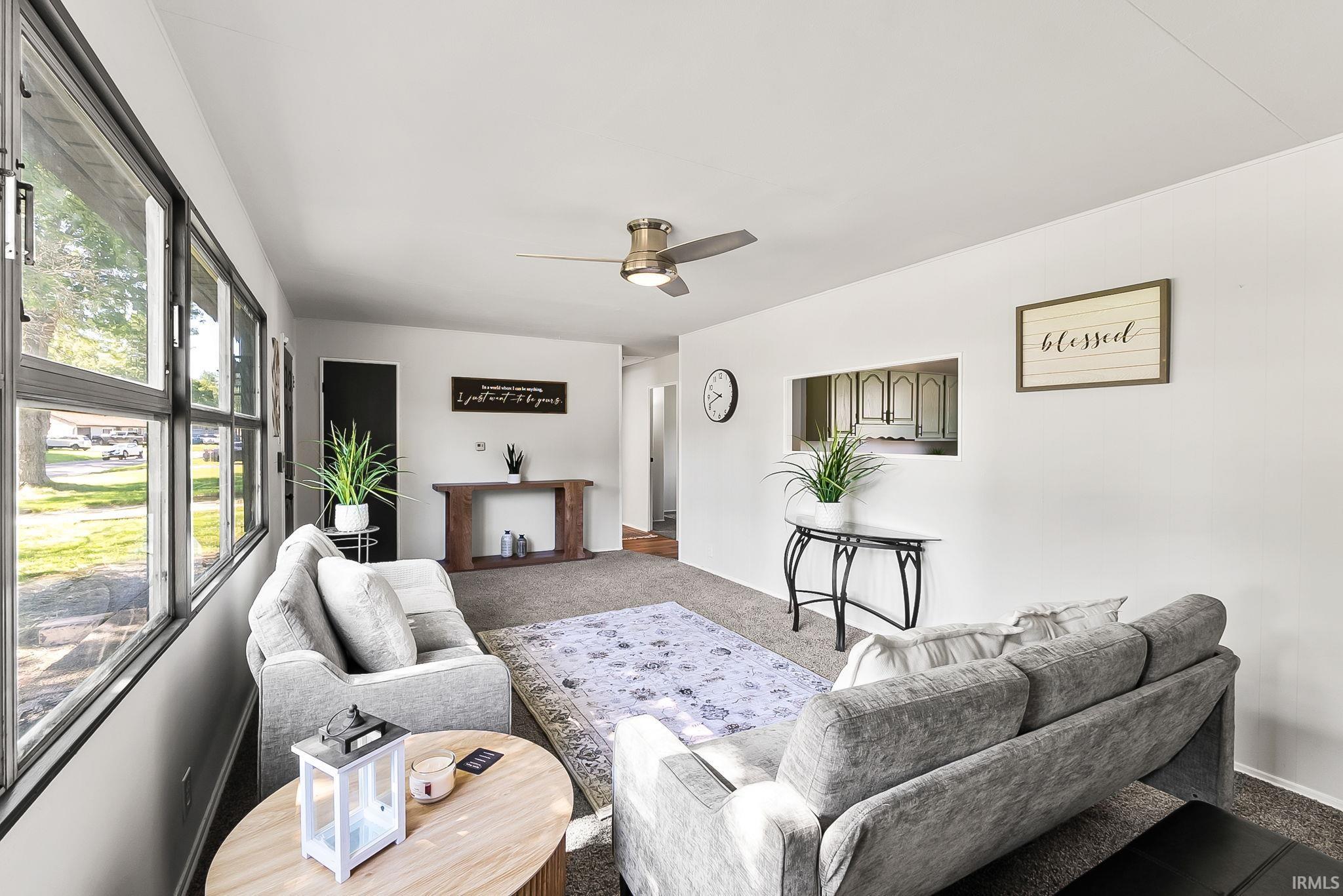 Bright living area featuring a wall of windows, neutral carpeting, a ceiling fan, a pass-through window to the kitchen, and a dark paneled door