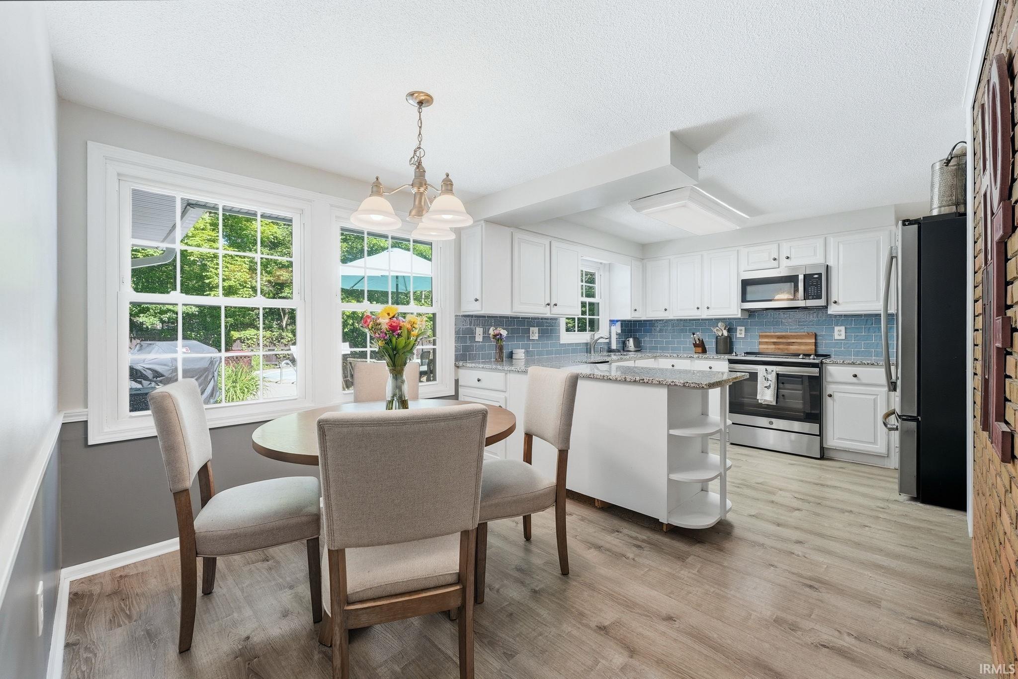 Open-concept kitchen and dining area featuring light wood-finish flooring, white cabinetry, a blue subway tile backsplash, stainless steel appliances, and a central island with granite countertop