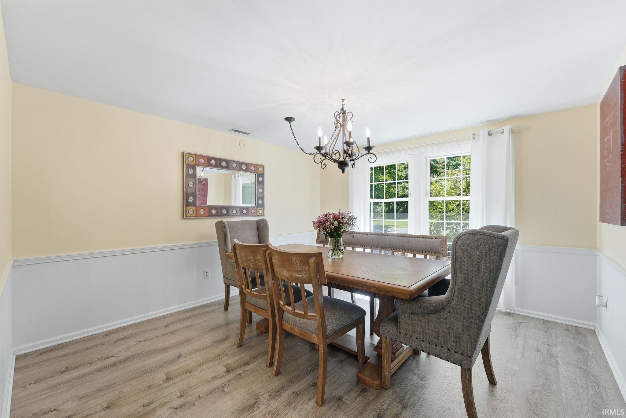 Dining area featuring wood-finish flooring, white wainscoting, an overhead chandelier, and double windows with grid patterns